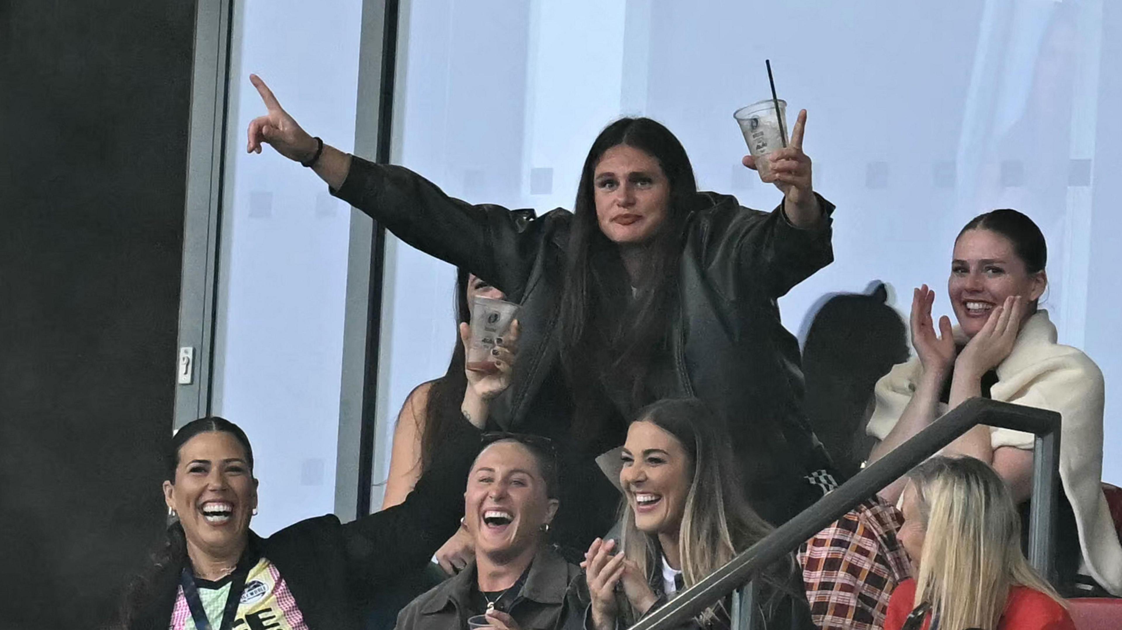 American rugby union player and social media star Ilona Maher stands with her arms wide, and is holding a drink with a straw in her left hand, as she reacts to her face being shown on the big screen at Ashton Gate. A small group of other women are laughing as they sit around her