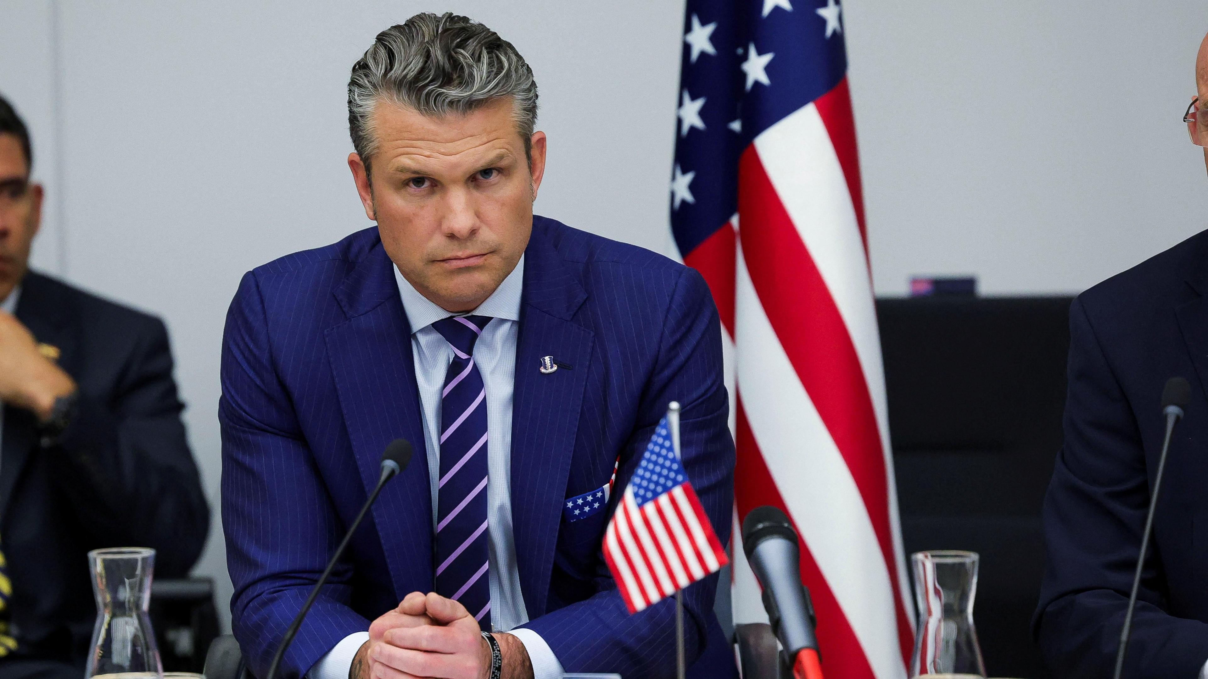 a seated Hegseth in blue pinstripe jacket with blue tie striped in pink and American flag handkerchief, leans forward toward a black microphone looking stern with a small American flag before him to his left on the table and a large US flag behind him, also to his left