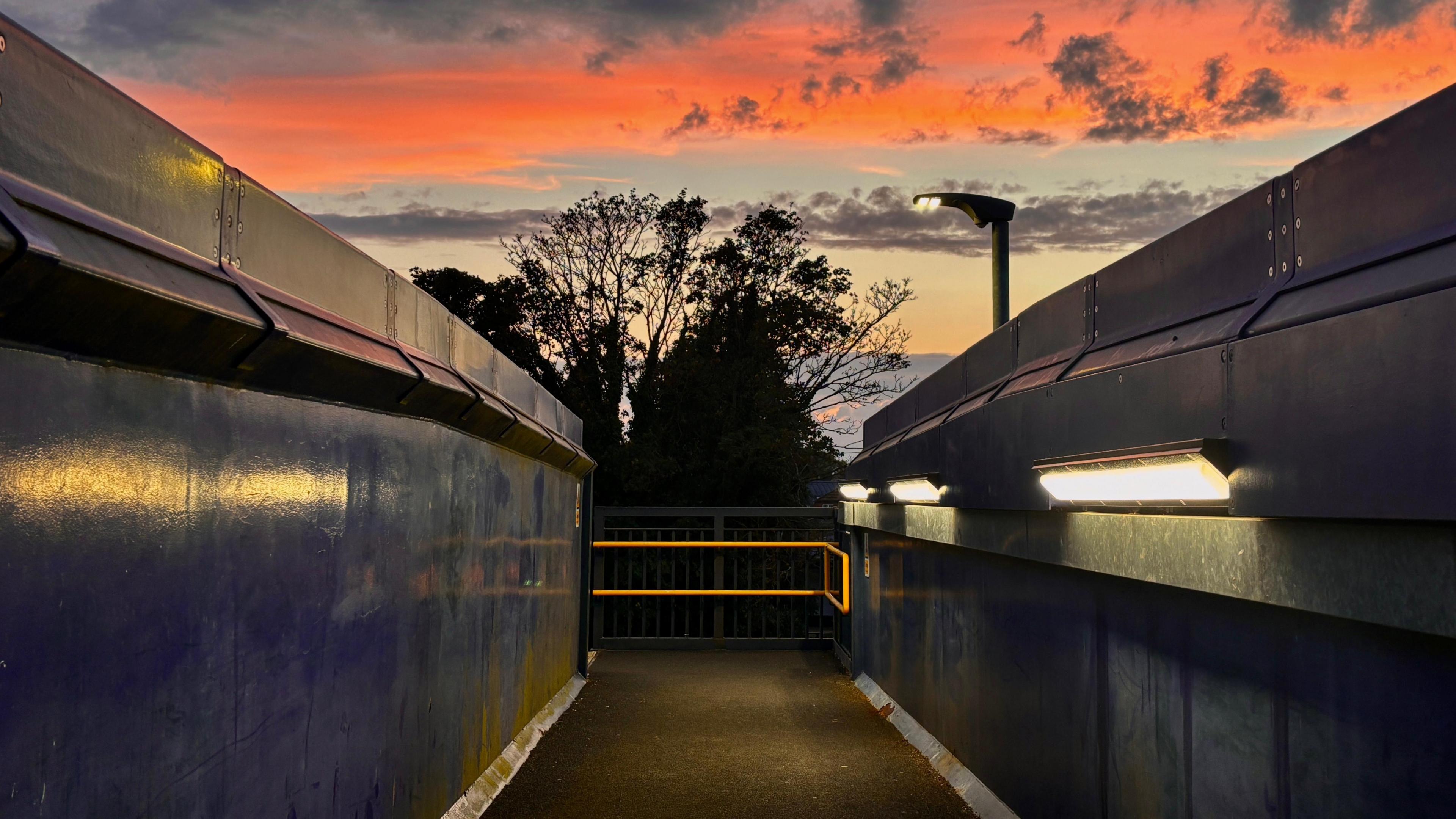 A beautiful skyline with wispy clouds and an orange glow above what appears to be a bridge walkway with lights on one side and a handrail at the far end.