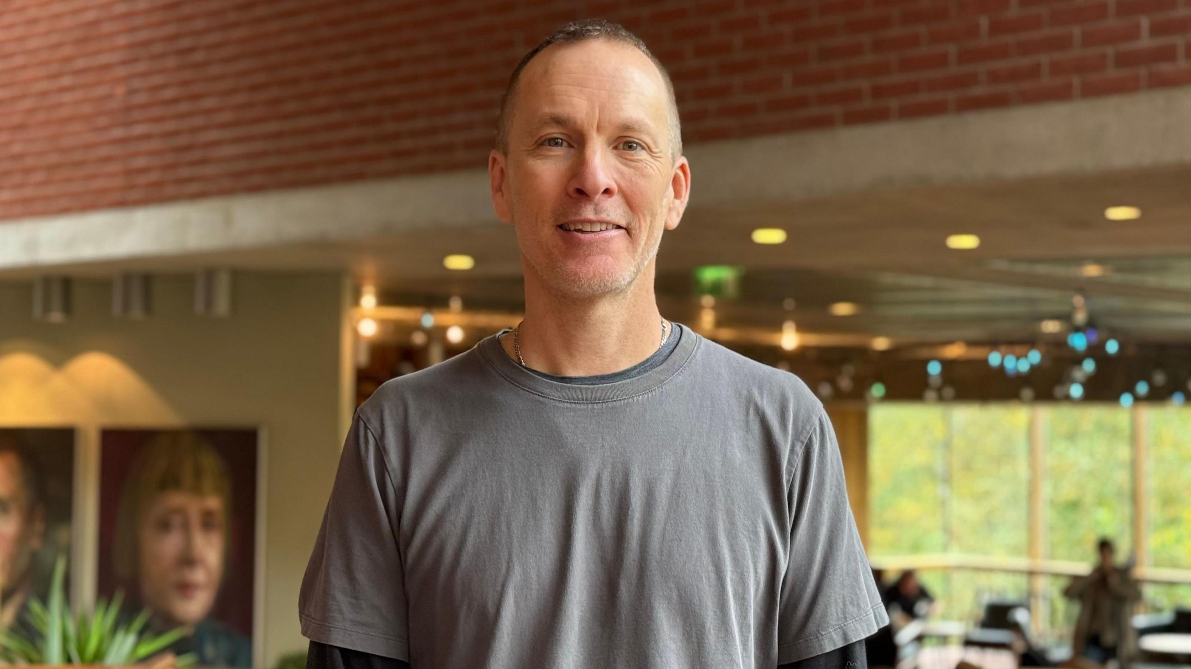 A man in a grey t-shirt smiles at the camera. He has short grey hair. There is a cafe behind her. There are large photos on the wall.