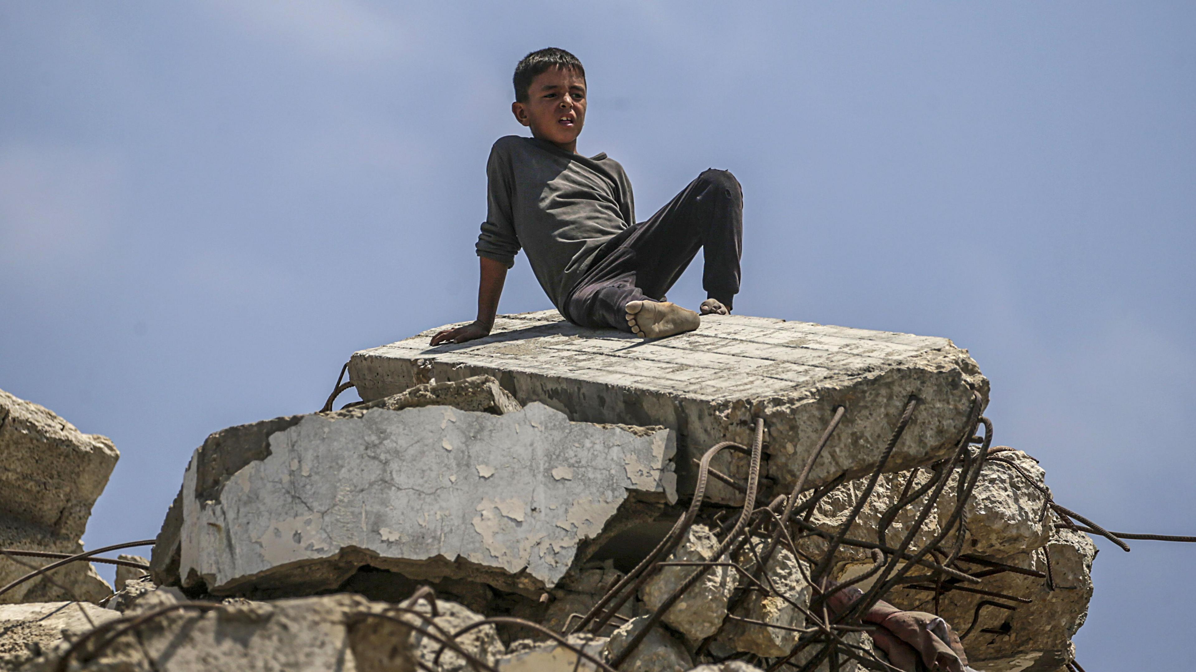 A young boy sits atop large chunks of rubble. Metal prongs poke out from the block he sits on. His feet are dirty and he wears dark trousers and a dark long-sleeve top, rolled up to the elbow on his right side. There is a blue sky behind him.