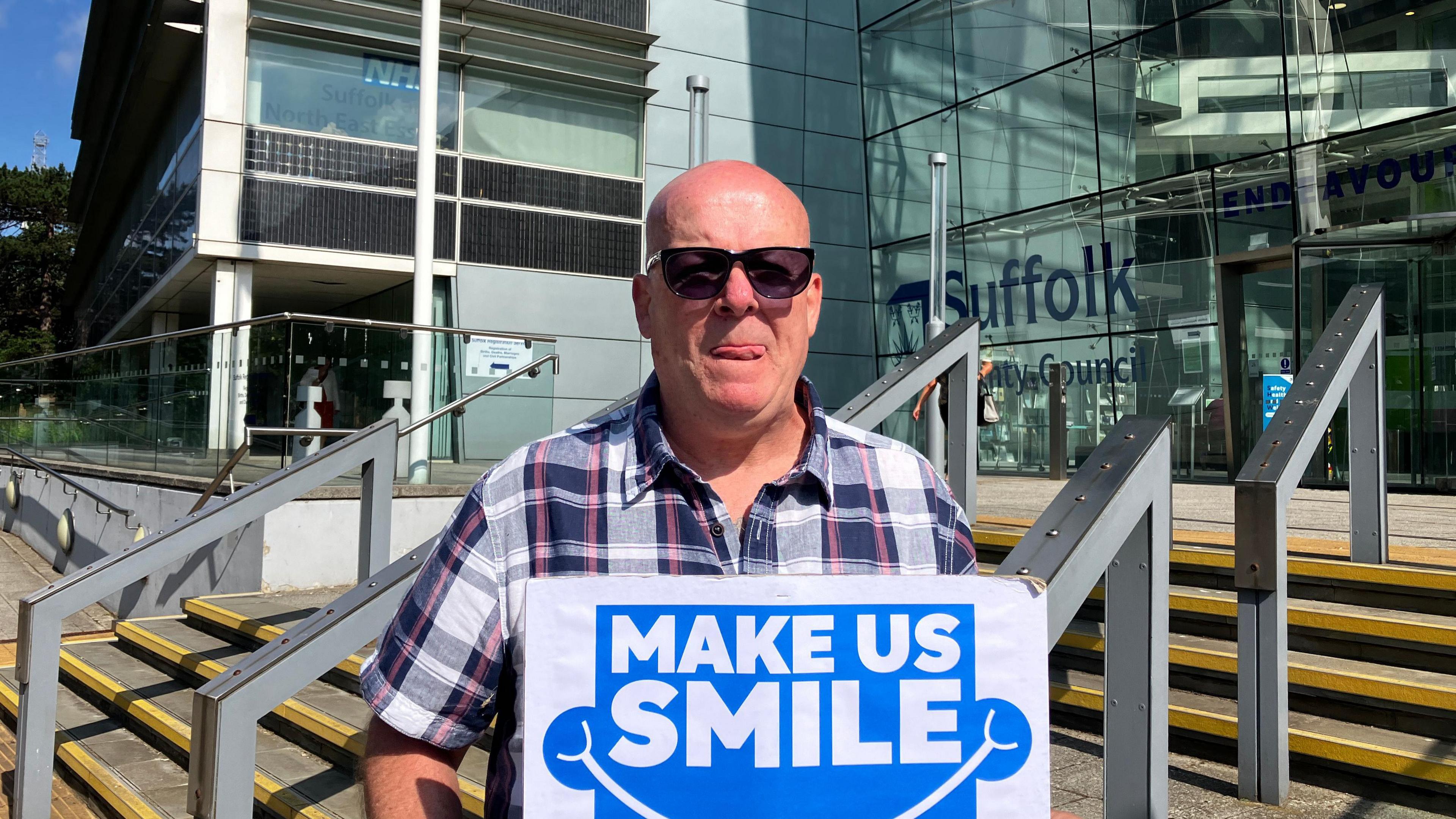 A bald man wearing sunglasses and a chequered shirt holding a placard which reads 'Make Us Smile'. 