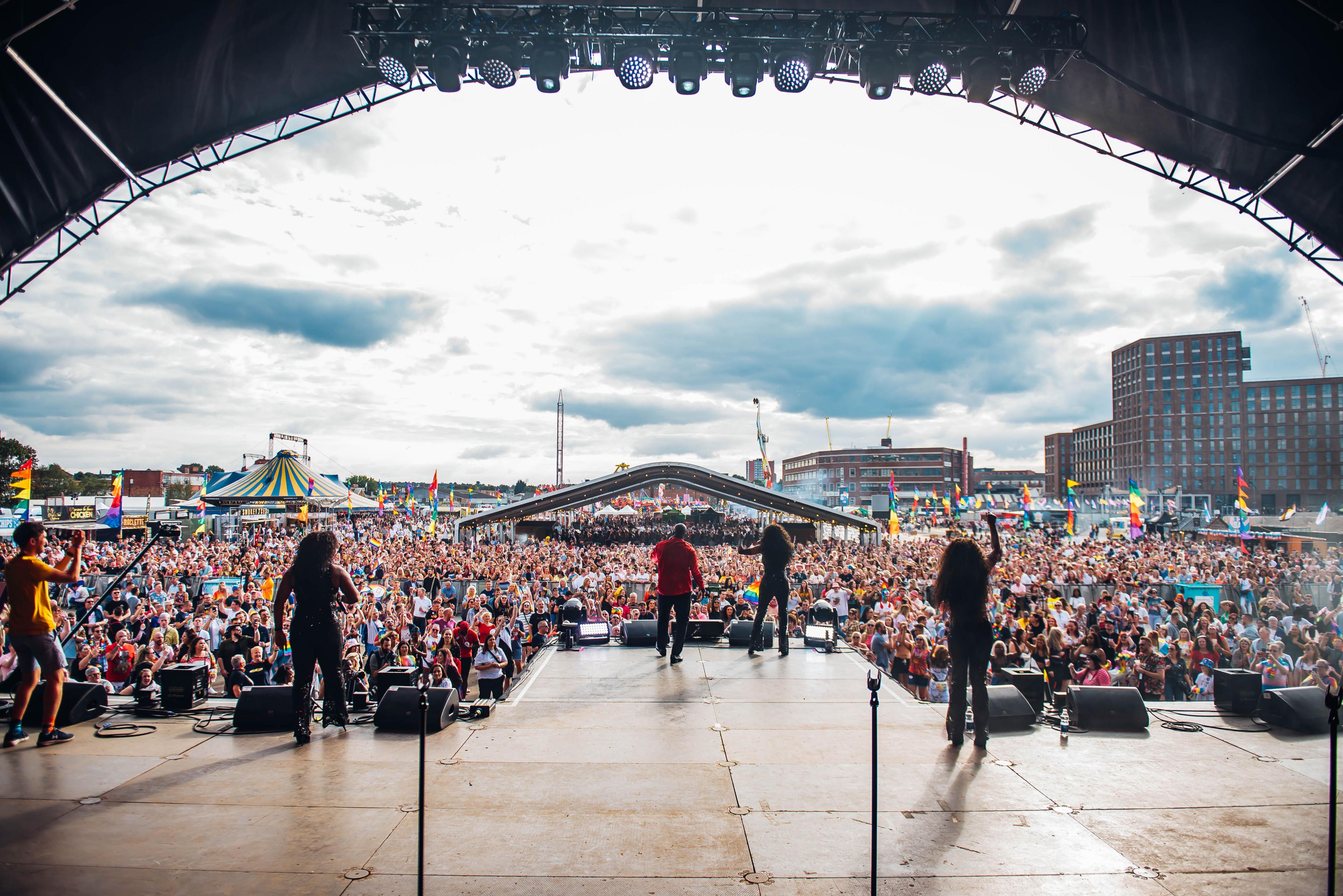 Thousands of people attending Birmingham Pride at Smithfield, as presenters introduce acts to the main stage. A sea of people leads off from a stage with tower blocks in the distance.