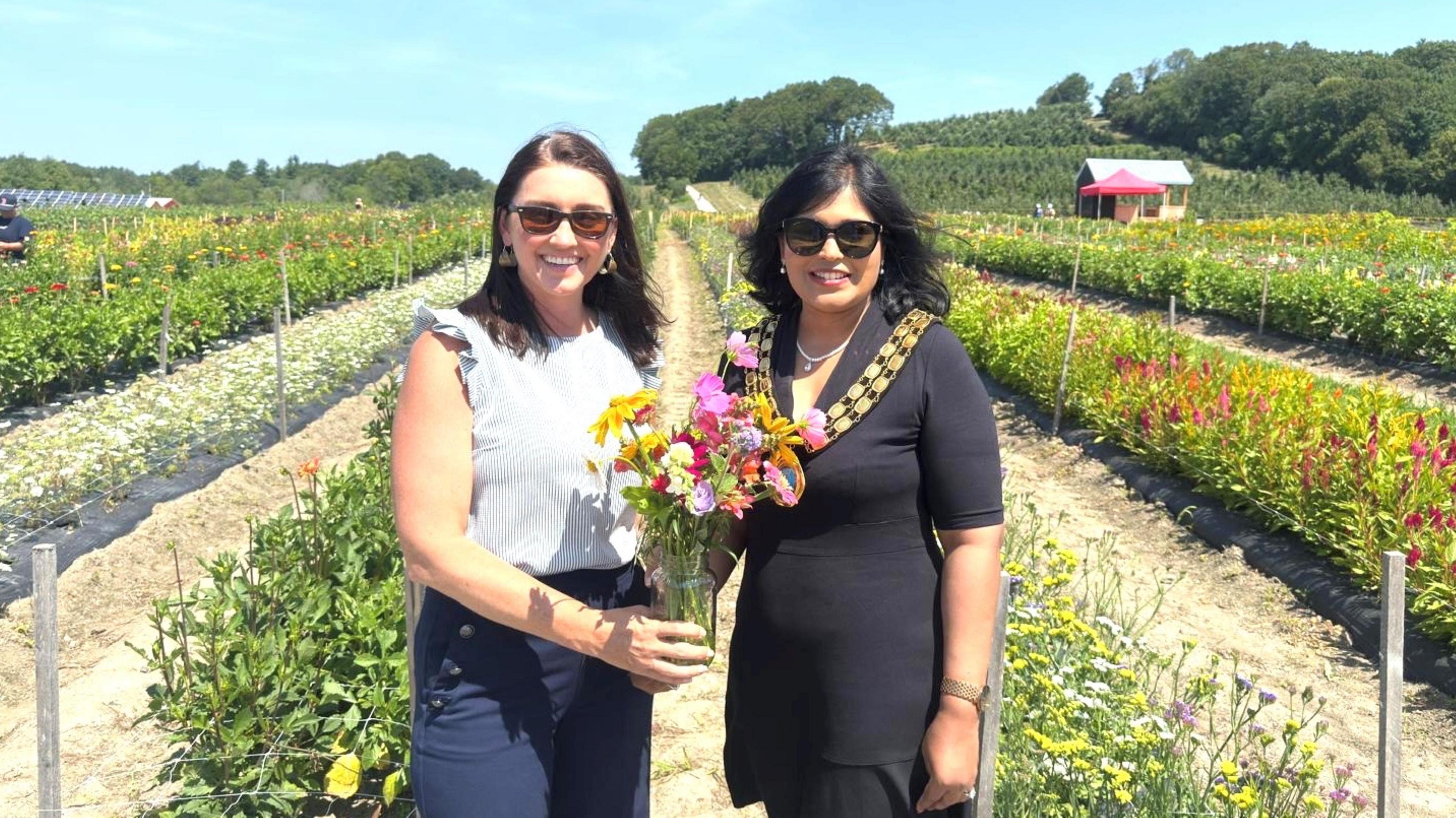Two women standing together in amongst rows of flowers. The woman on the left is wearing a white sleeveless blouse and blue trousers. The woman on the right is wearing a black dress, a mayoral chain and the two of them are holding a small bouquet of flowers.