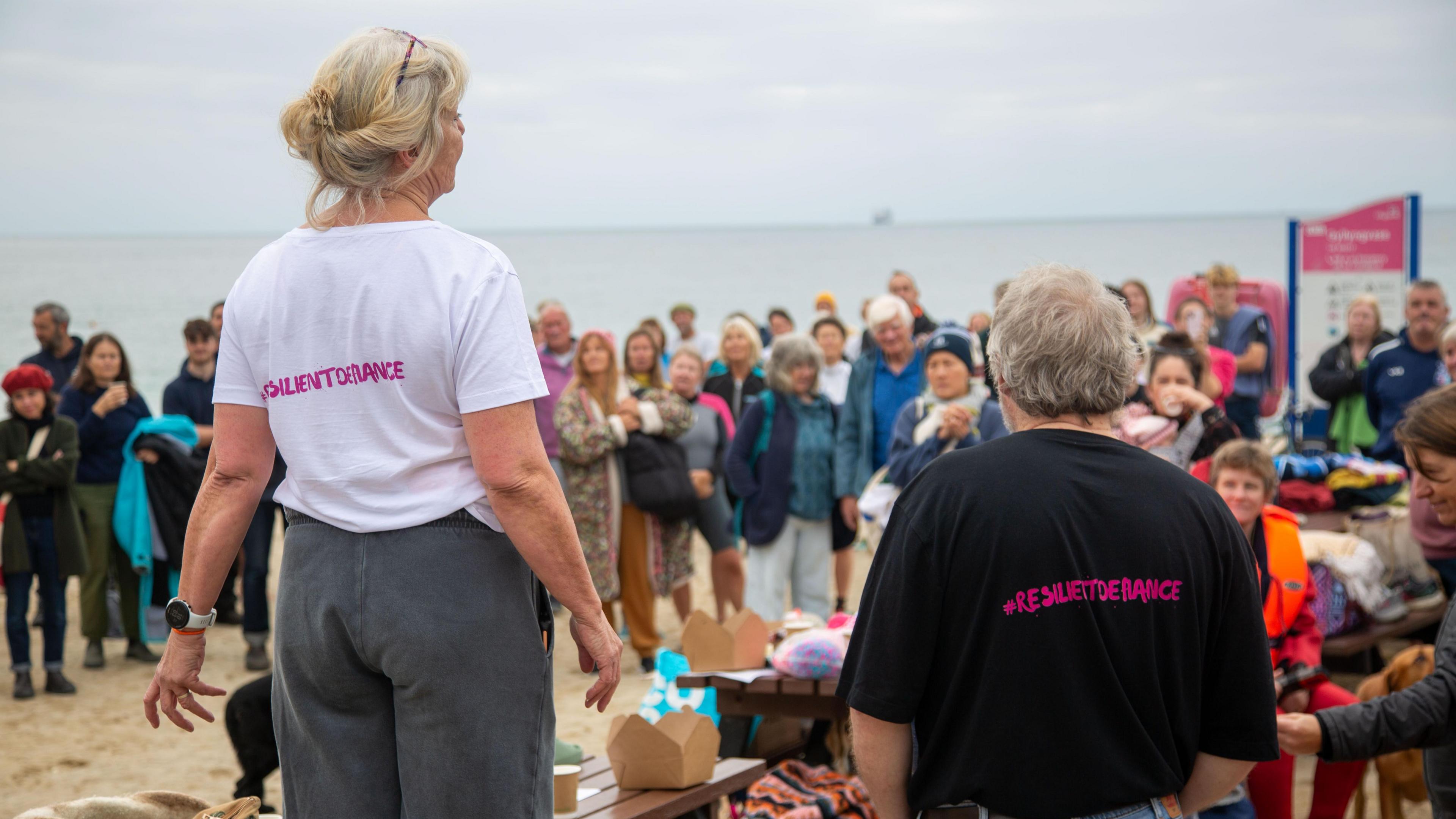 Ruth Hitchcock and Dave Hitchcock with their backs to the camera and facing a crowd of people. They are wearing t-shirts which say #resilientdefiance on the back in pink letters. The sky is grey. The sea is a light grey.