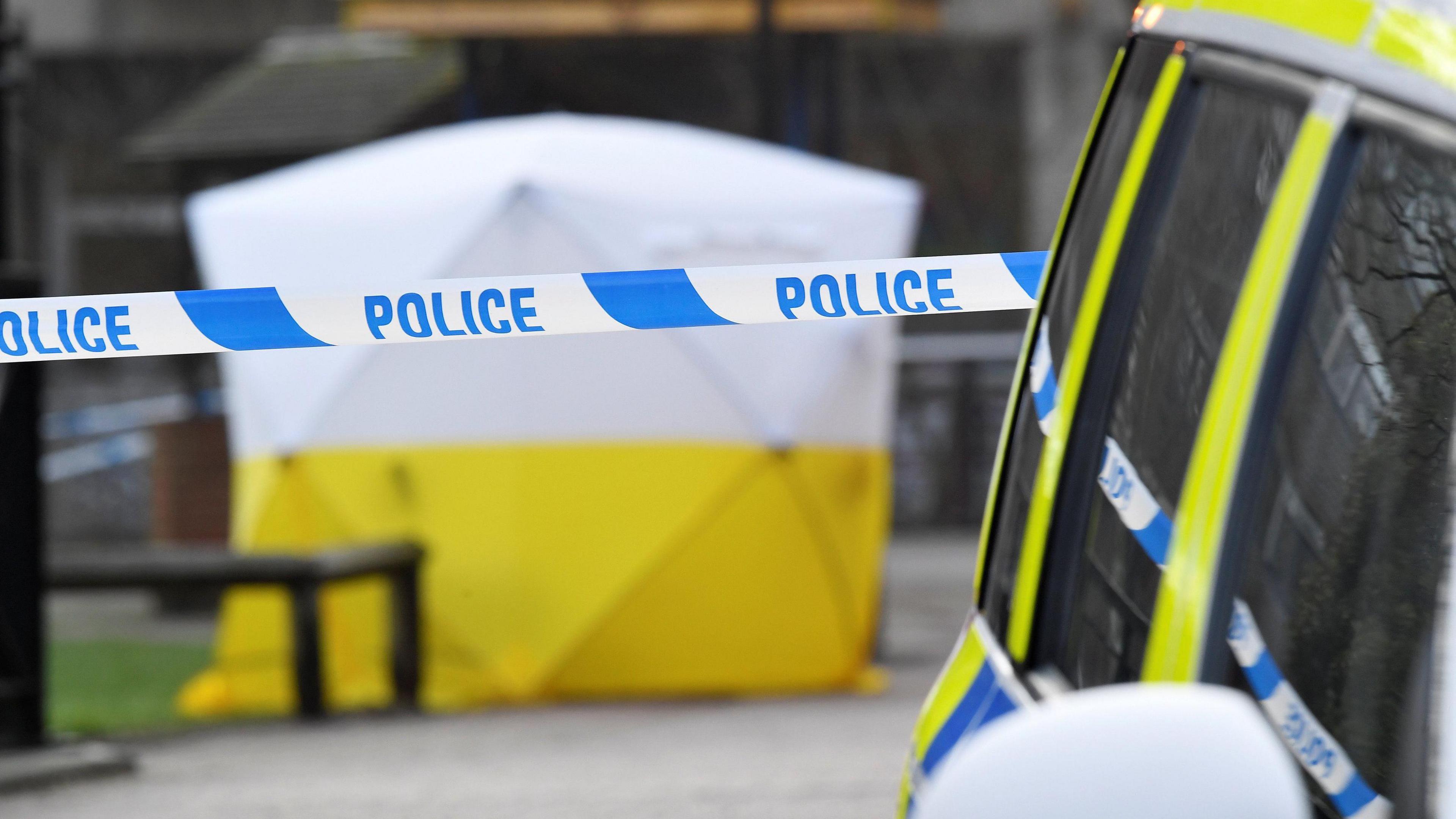 A police cordon, with a white and yellow tent in the background. The tent is covering the bench where Sergei and Yulia Skripal were found. 