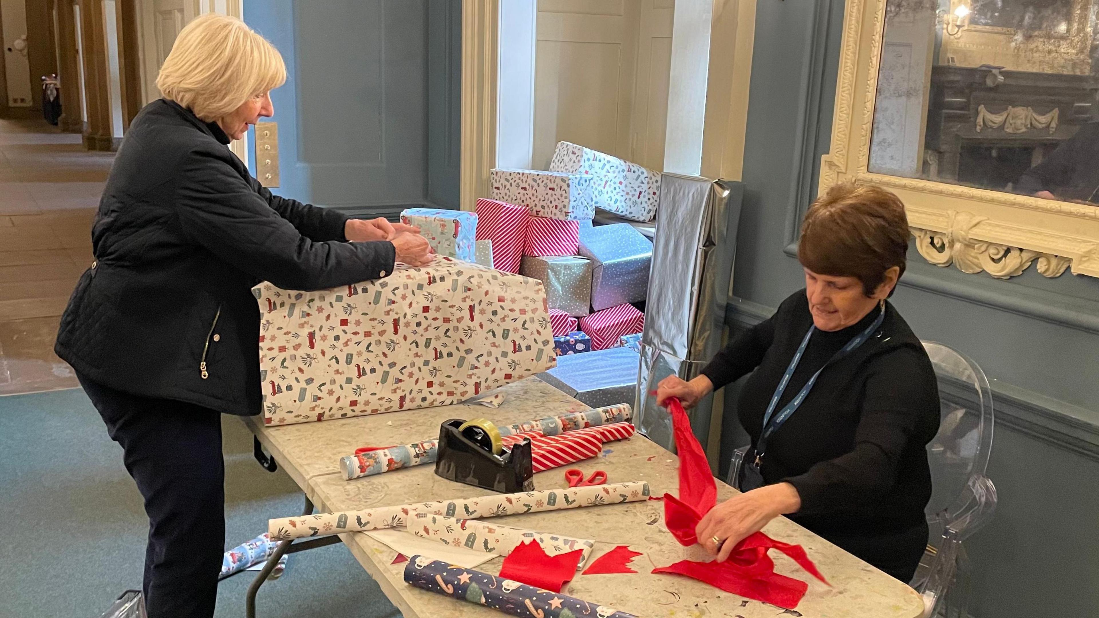 Two volunteers are wrapping boxes in Christmas paper and creating bows out of ribbon.