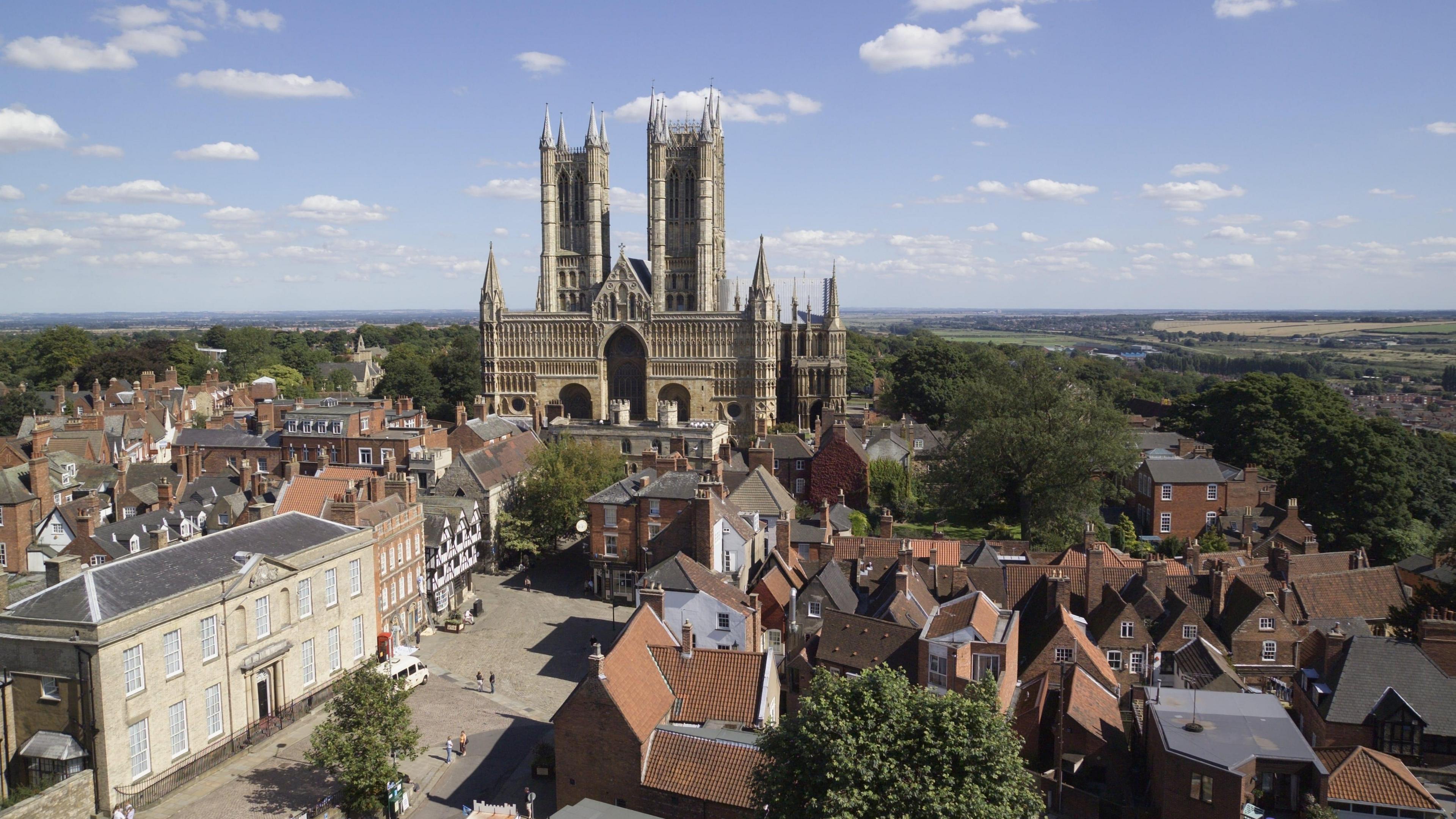 An aerial view of Lincoln Cathedral with its tall towers and intricate stone architecture. The cathedral is surrounded by historic red-brick buildings with pitched roofs, narrow streets, and a small square in the foreground. Green trees are also visible. The background stretches into open countryside under a blue sky dotted with clouds.