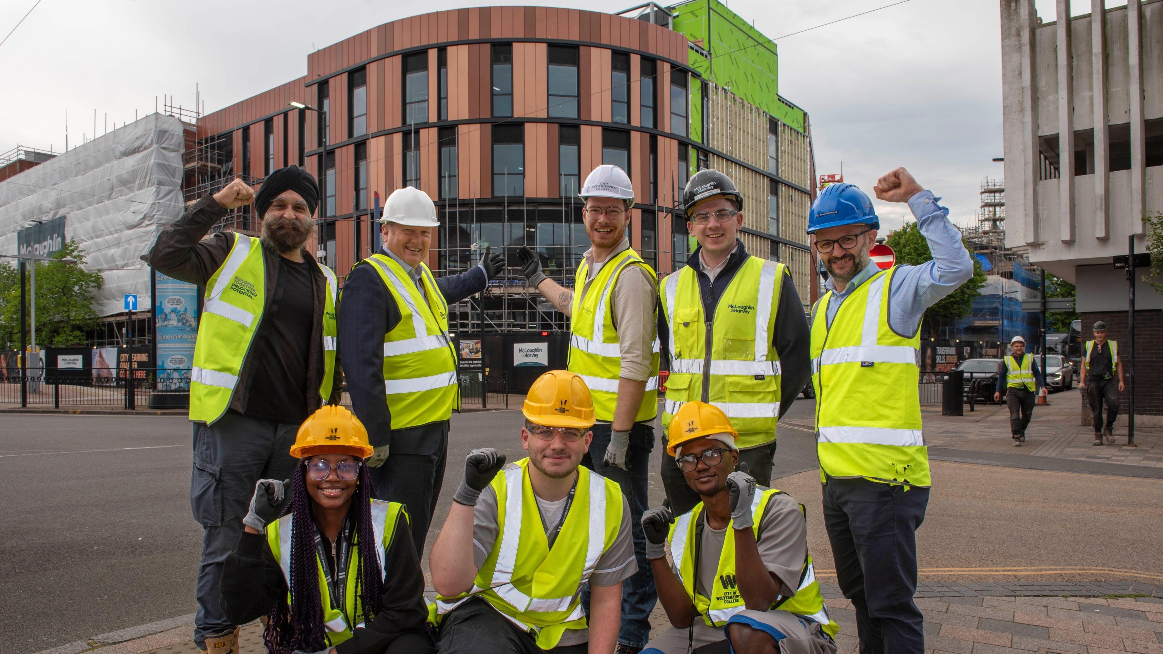 A group of people wearing helmets and hi-vis jackets stand in front of the new college campus. The building has terracotta coloured cladding. Some protective covering and scaffolding can be seen on the exterior. Street signs and boards can be seen in front of the building.