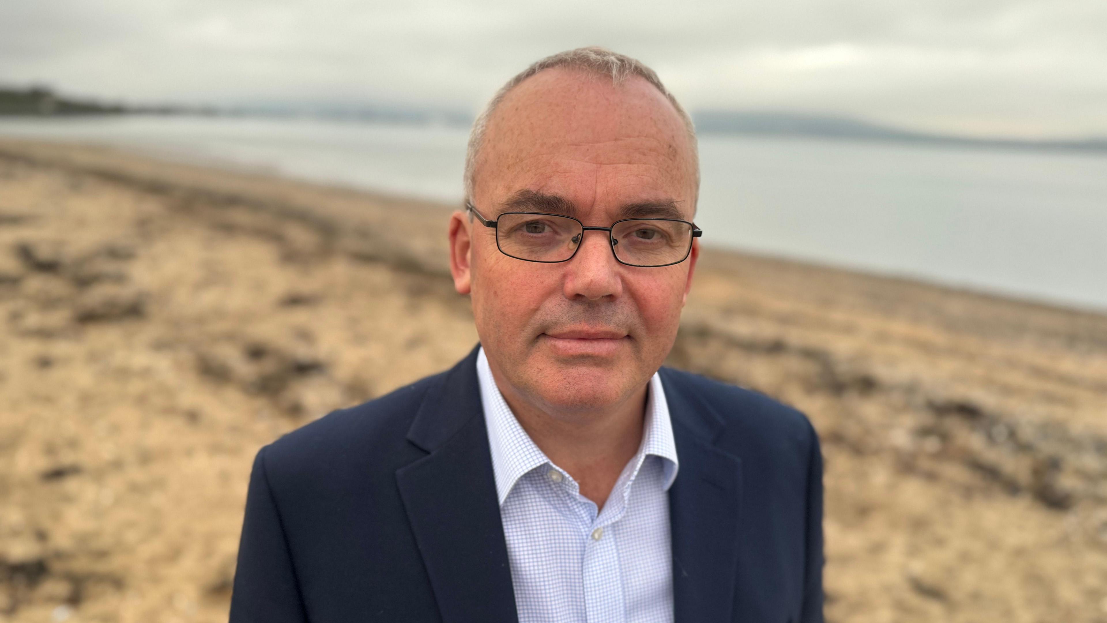 A man stood on the beach in a navy blue suit, he is wearing spectacles. 