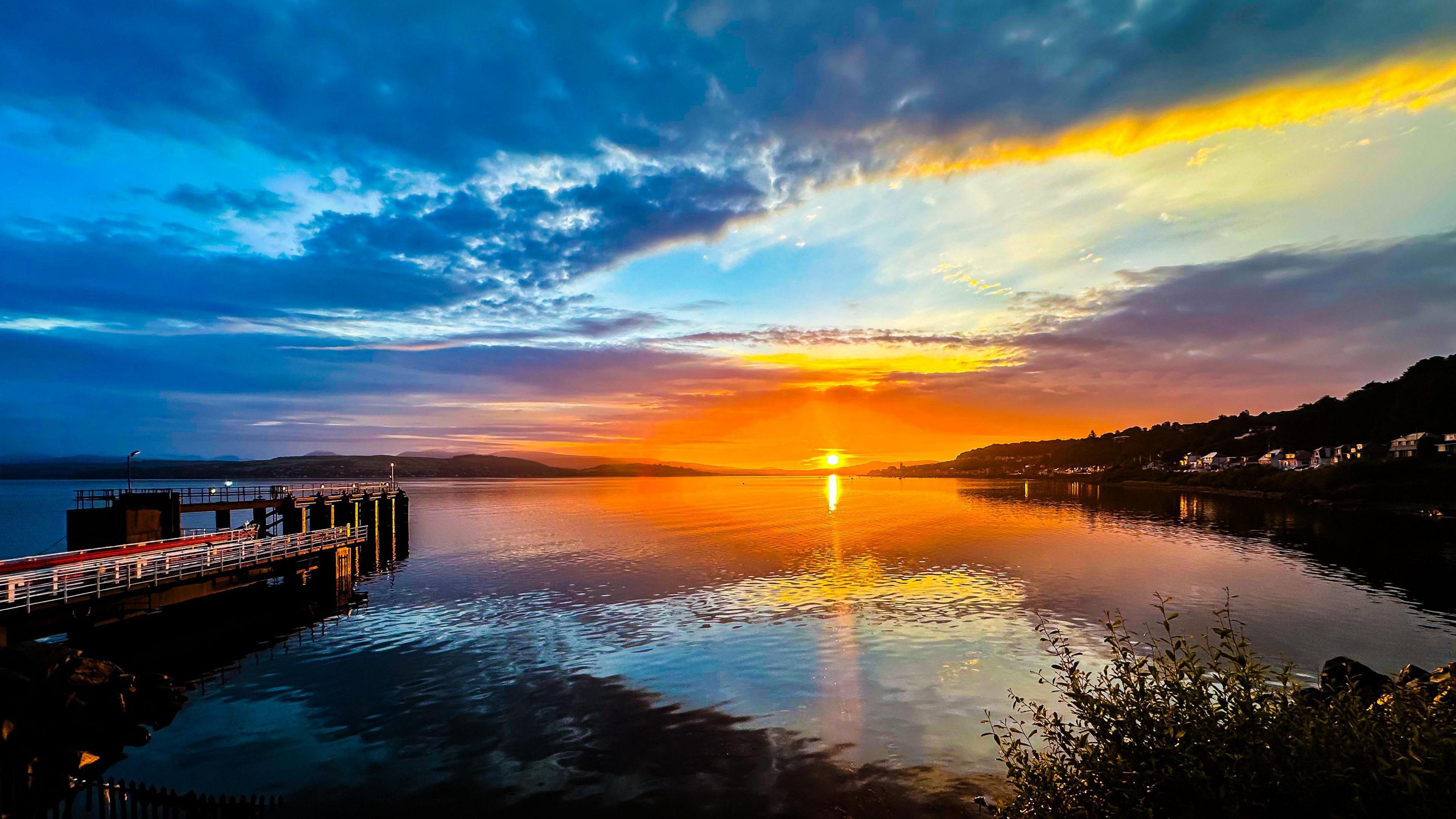 An orange sunrise over a calm body of water. The sun is just rising above the horizon and the sky is a mixture of blue, grey and orange. The water is still and calm and is reflecting parts of the sky. A metal and stone pier stretches out into the water on the right hand side.