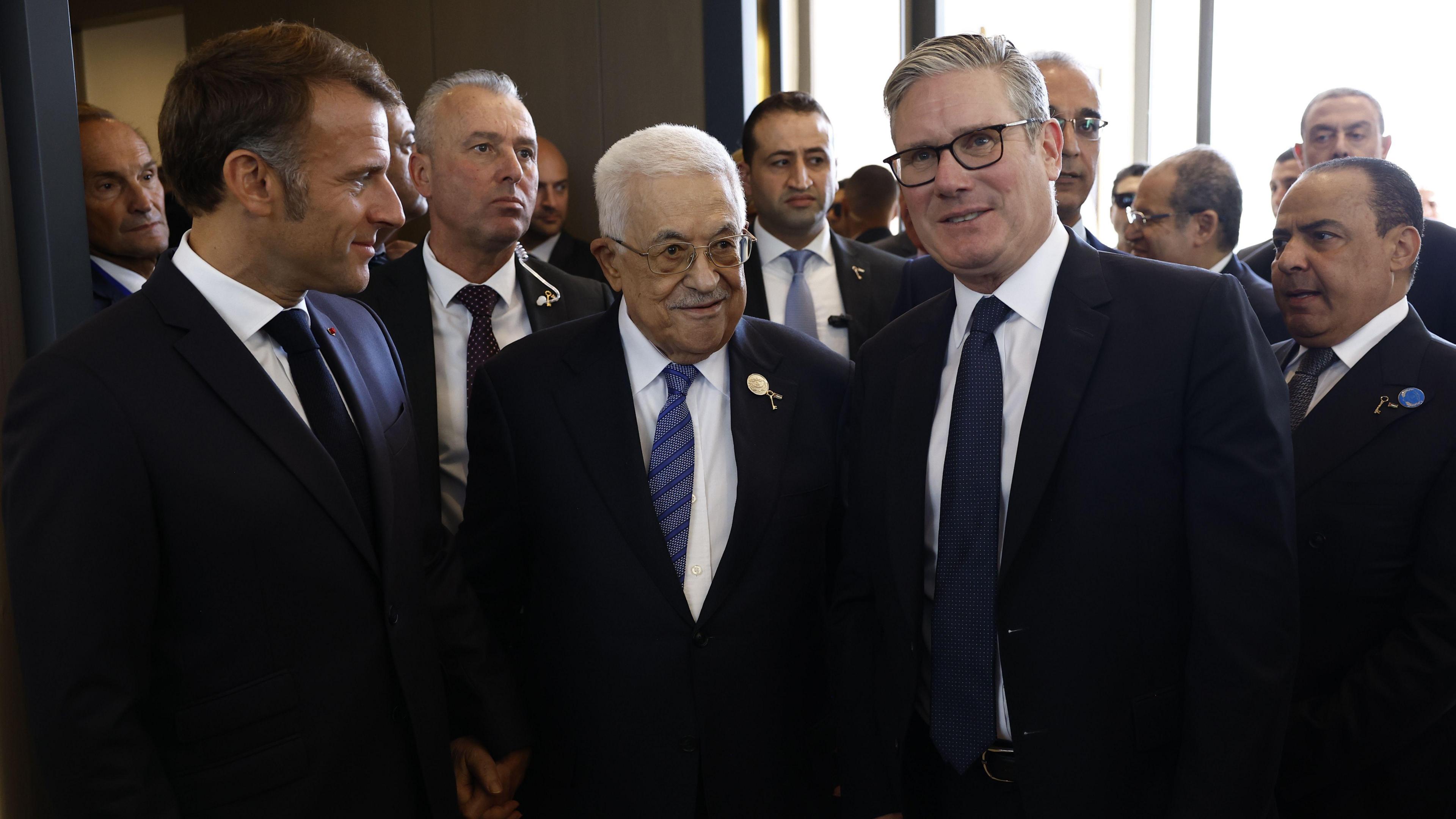 French President Emmanuel Macron (left) and British Prime Minister Keir (right) Starmer stand with President of the State of Palestine Mahmoud Abbas (centre). They are all wearing dark coloured suits.