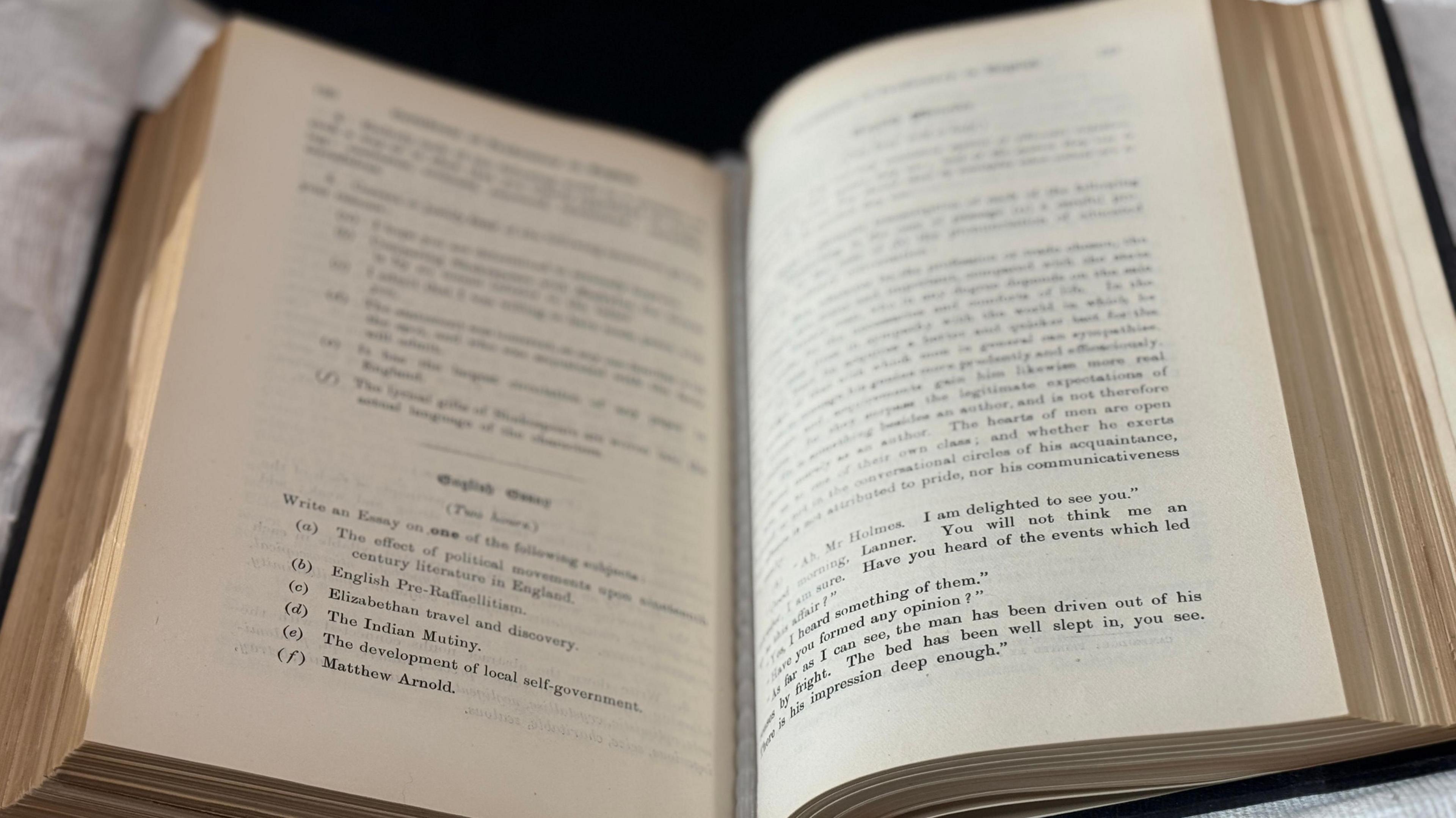 A book is displayed on a protective cushion. It is open, showing some of the questions from the first Cambridge English exam in 1913.