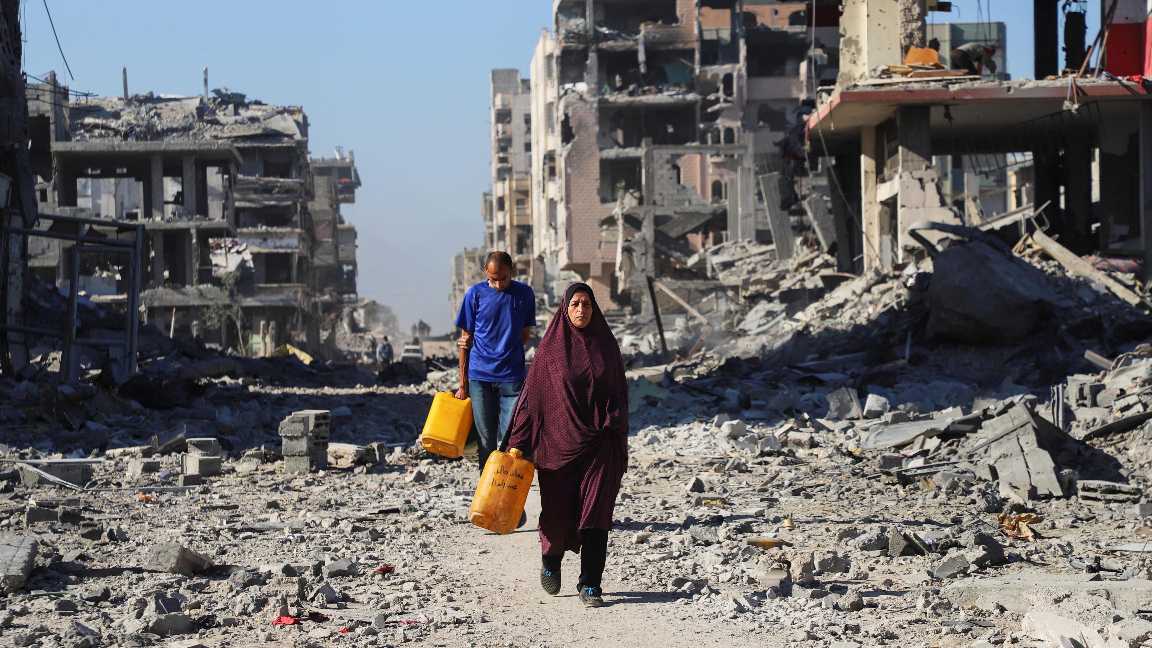 A woman in a maroon skirt and headscarf walks through rubble, carrying a yellow container. Behind her, a man in a blue T-shirt holds a similar container.