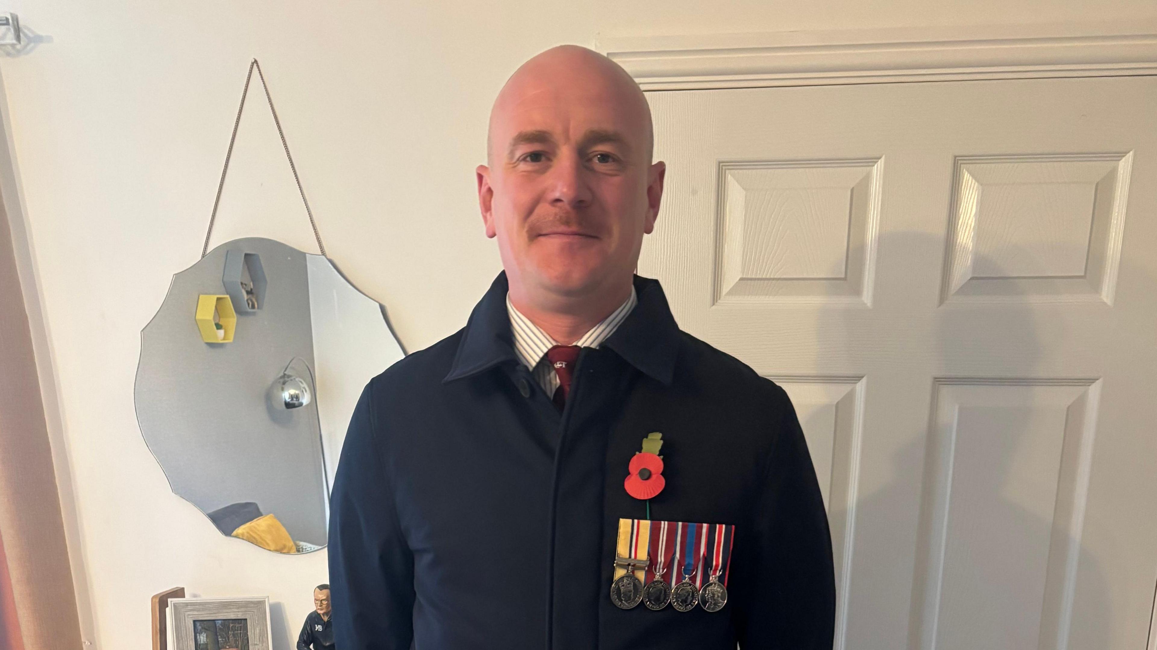 A man wearing a smart navy coat with military medals and a red poppy. He is standing in a living room looking straight at the camera and smiling.