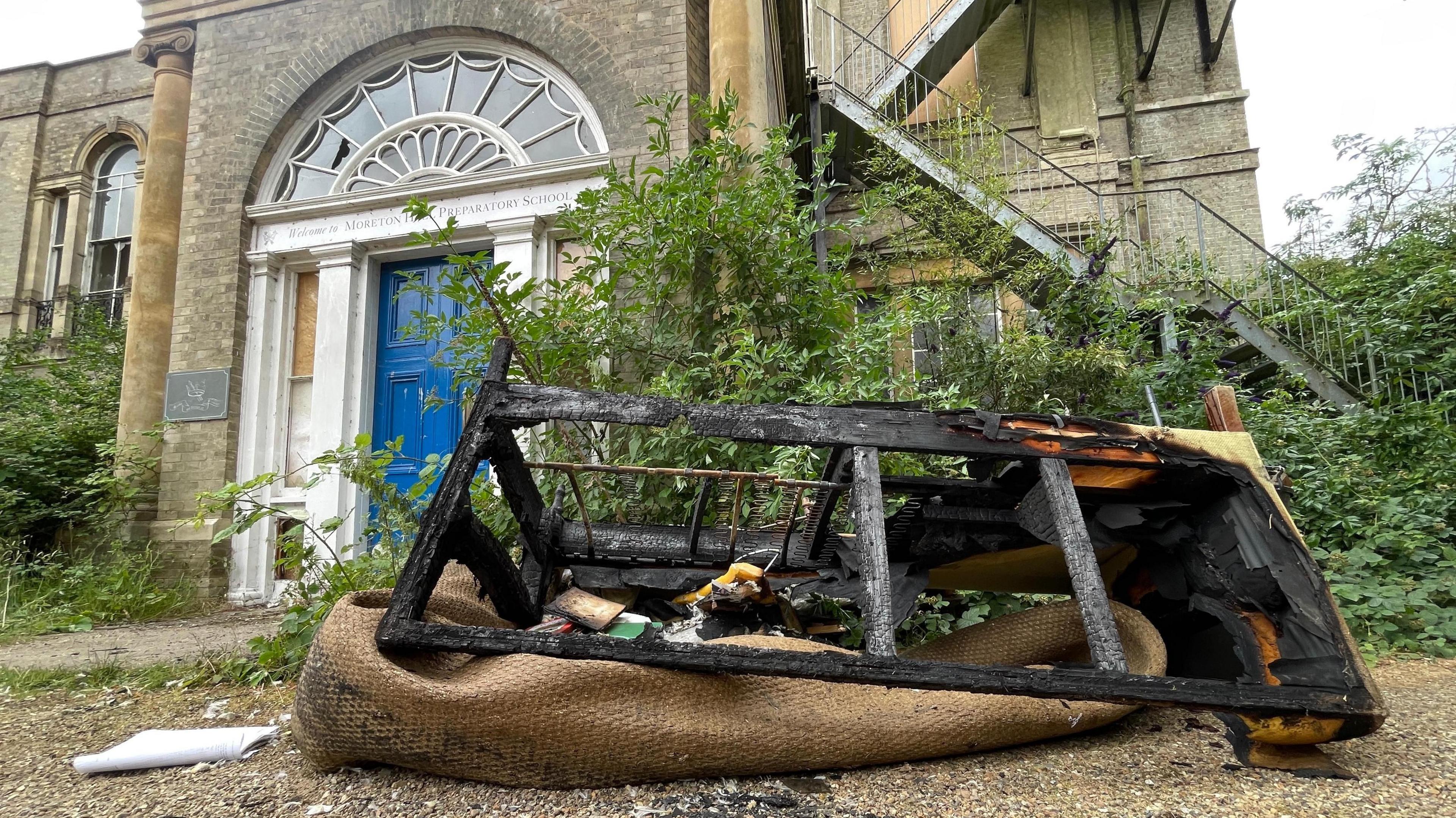 A burnt out sofa frame in front of Moreton Hall School. The blackened frame is resting on a piled up brown possible rug and there is other burnt debris resting on the rug. Behind the debris is the portico-style front door of the Georgian-style manor house. There are a lot of weeds and overgrown plants on either side of the front door.