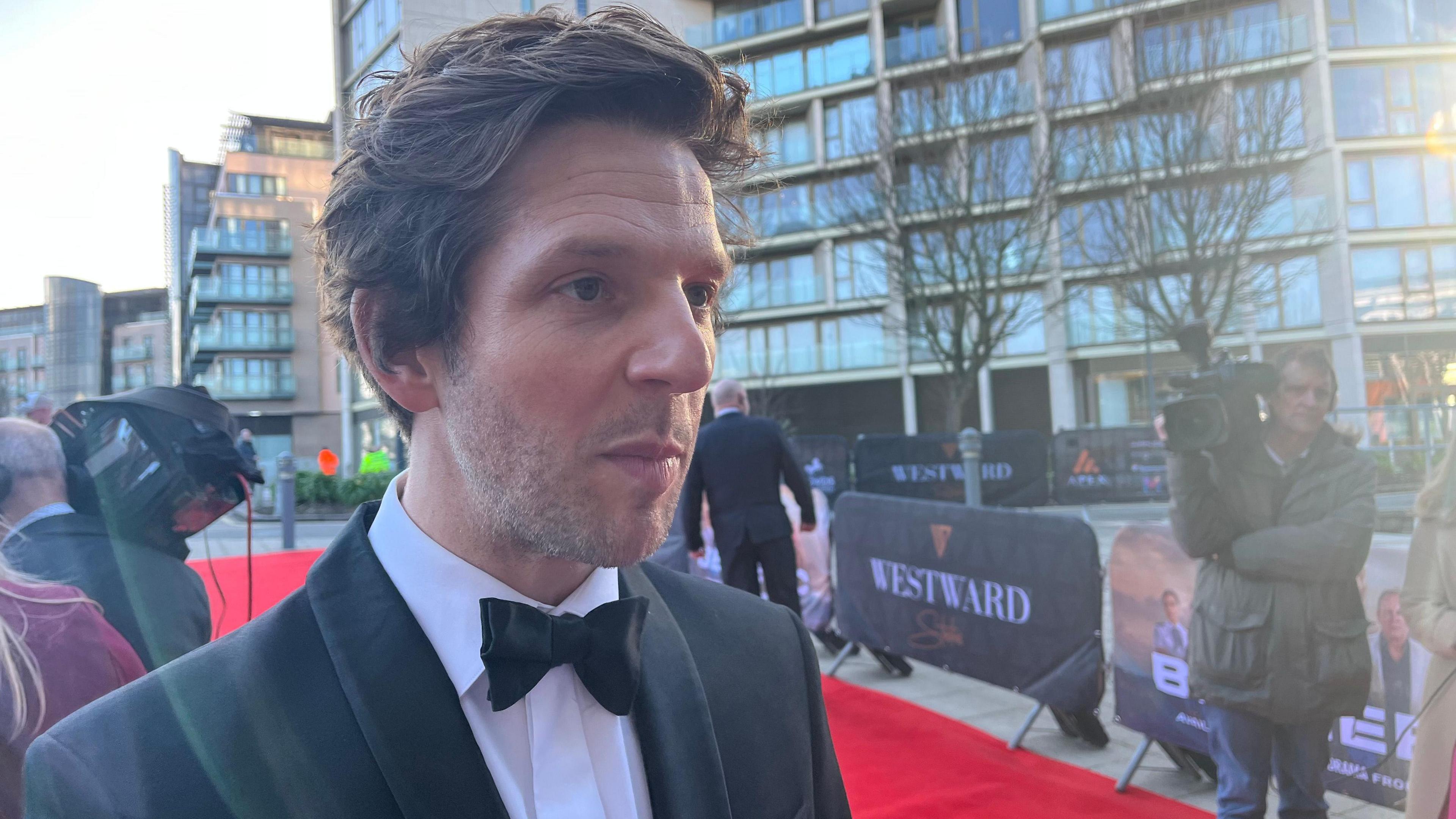 Damien Molony is being interviewed on the red carpet. He is wearing black tie. He has mid-length dark brown hair and a slight beard and moustache.