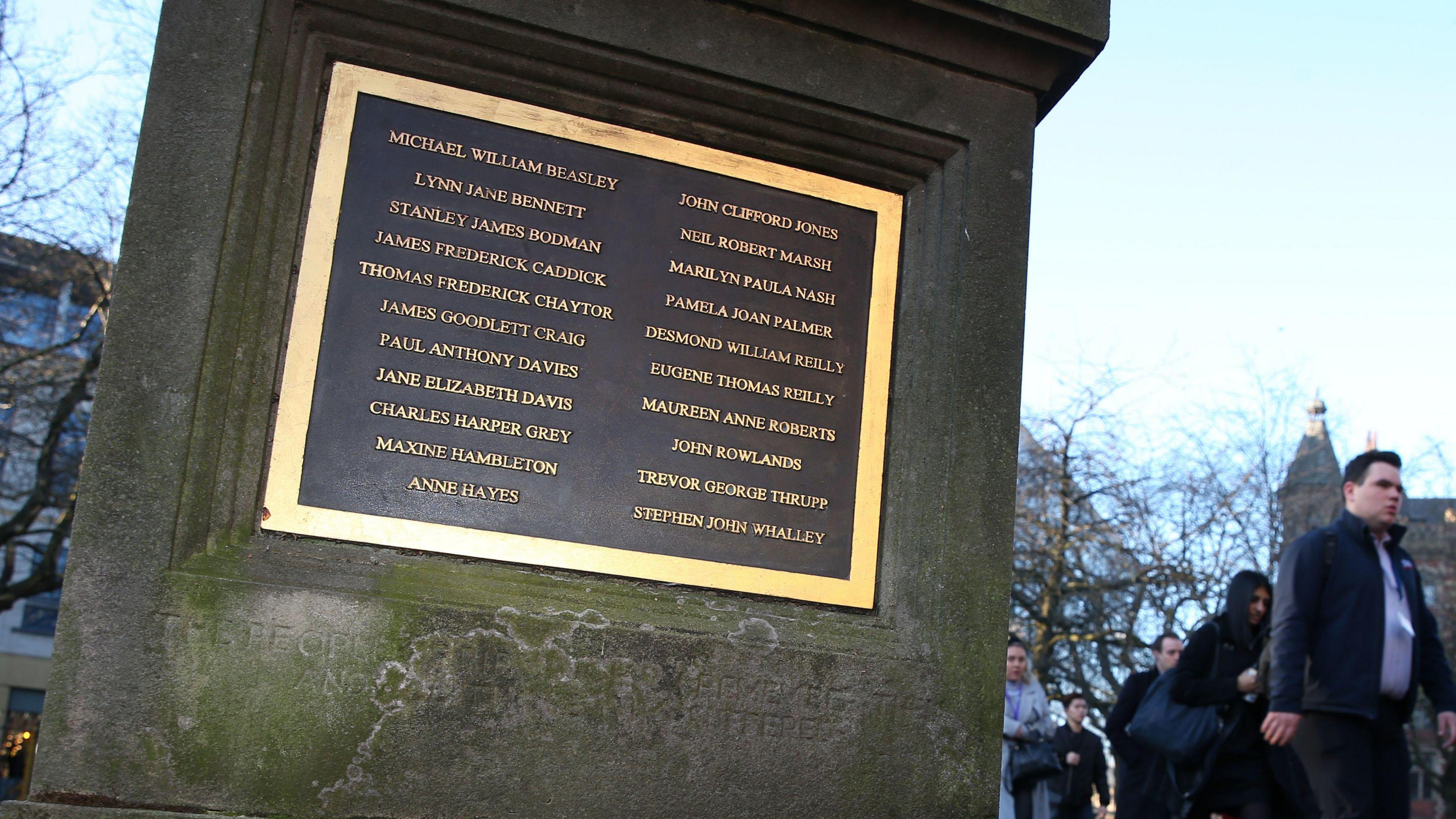 A stone plinth located outside in a tree-lined square, with a gold-rimmed black plaque that features 21 names in gold lettering. The general public can be seen on the right walking past the sign.