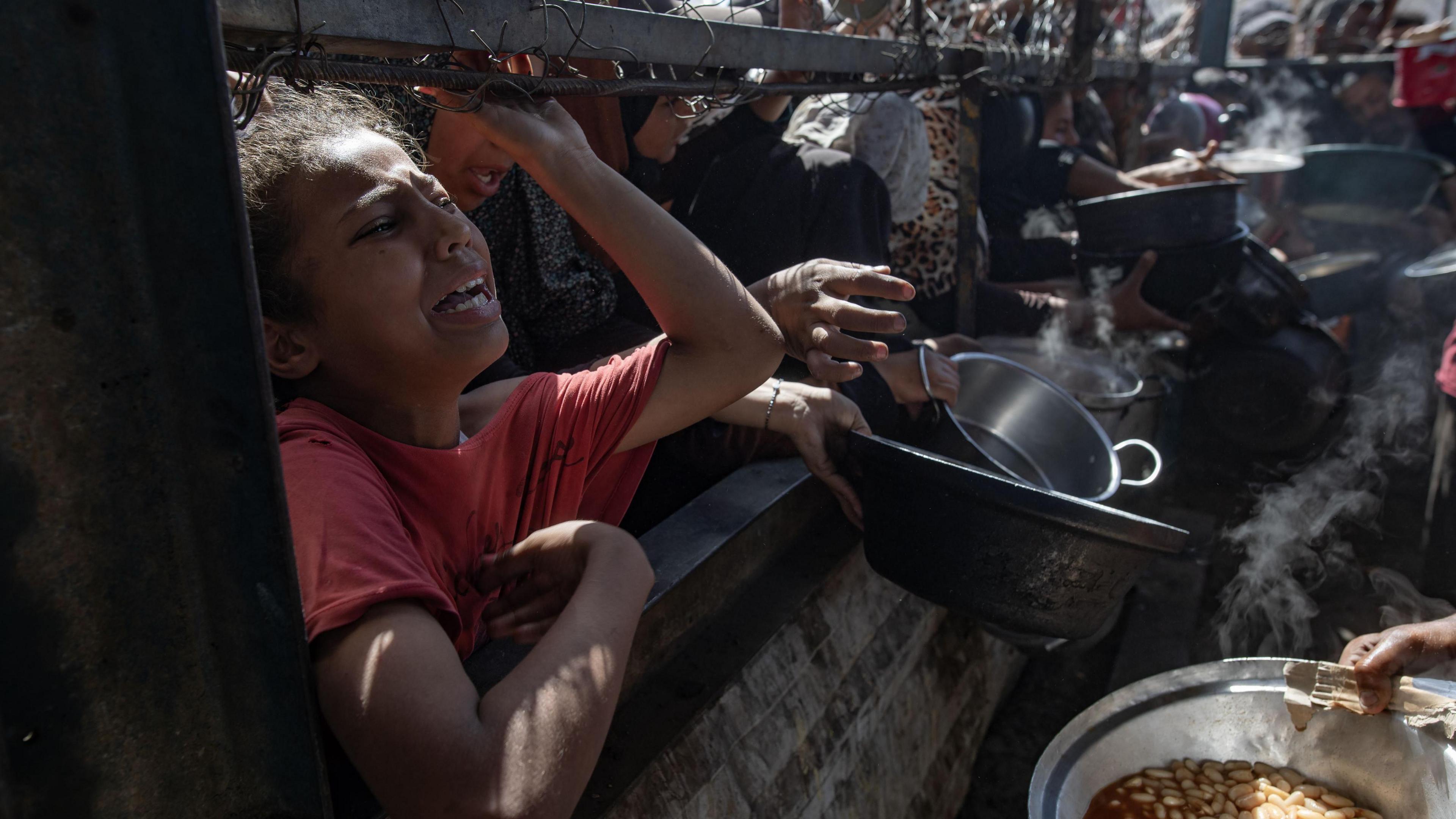 A girl cries out as she leans against the wall of a charity kitchen, beside people holding out empty pots and pans to be filled from a bowl of soup-like food