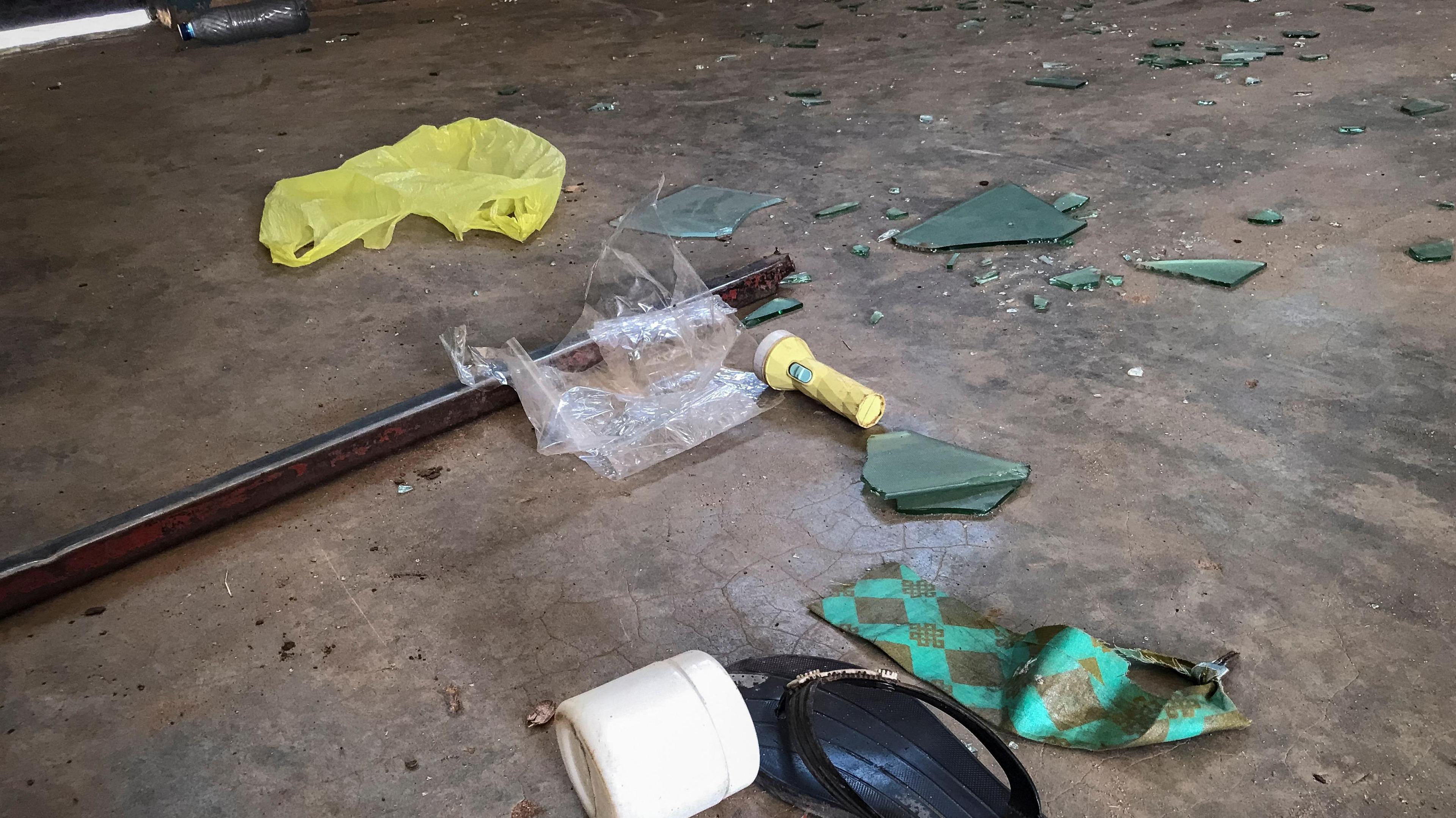 Scattered belongings on a floor inside a student dormitory. The ground is bare. Broken glass, a flip flop, a small torch and a plastic bag are on the floor. 