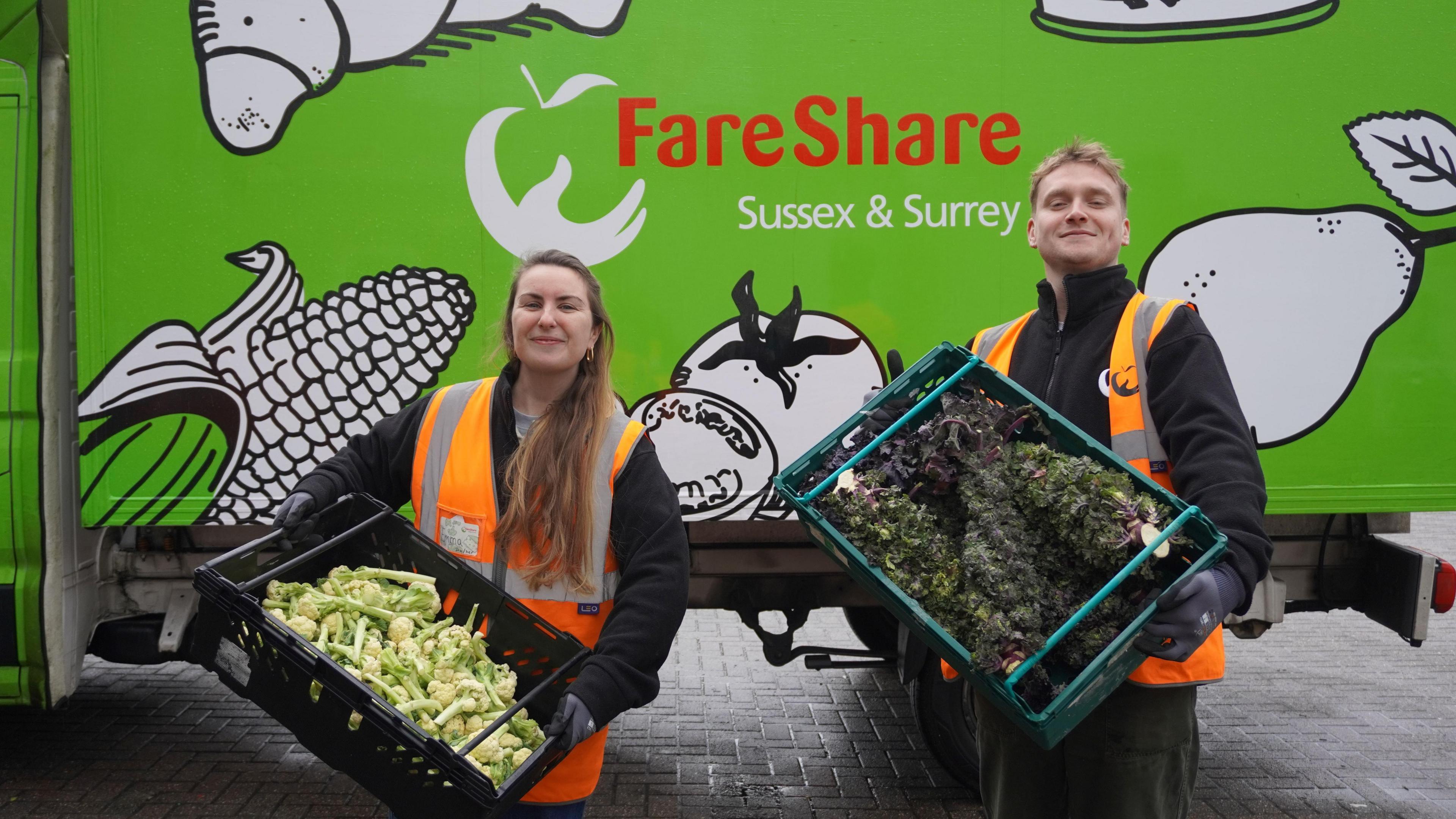 Two individuals stood in front of a bright green van which reads 'FareShare Sussex and Surrey'. They are both stood with crates of vegetables. On the left a woman with long brown hair and on the right a man with short blonde hair. They are both wearing orange Hi-Viz jackets. 