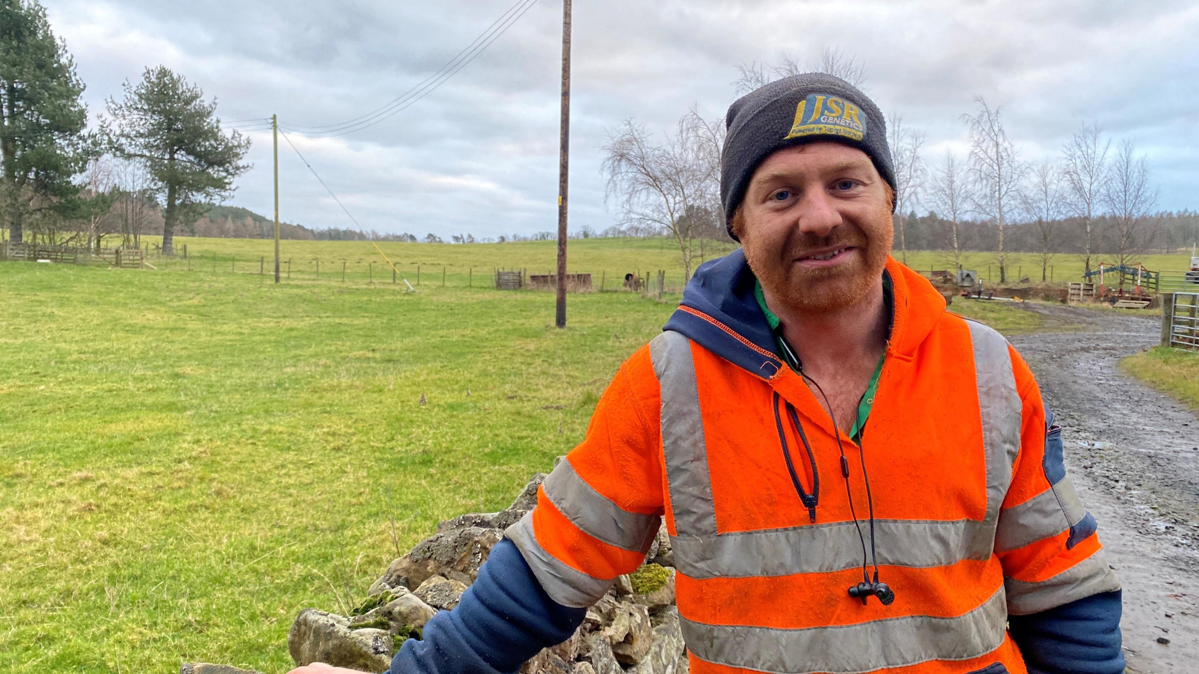 Ben Leyland is standing by a drystone wall. He's a 30-year-old man wearing a high vis orange jacket over a blue hoodie. He has a ginger beard and a grey hat 