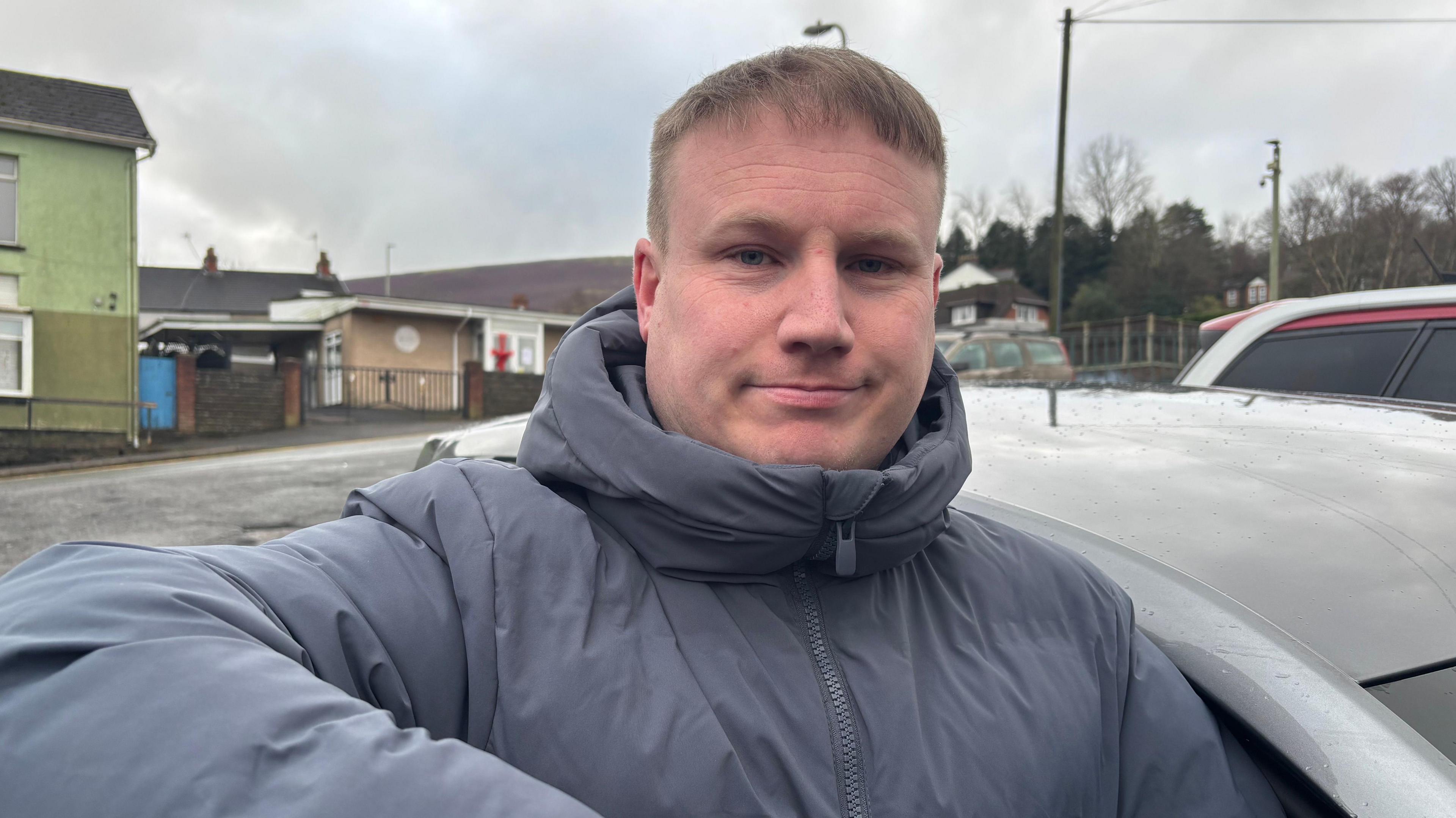 Dyn a chot glas yn sefyll wrth ei gar.