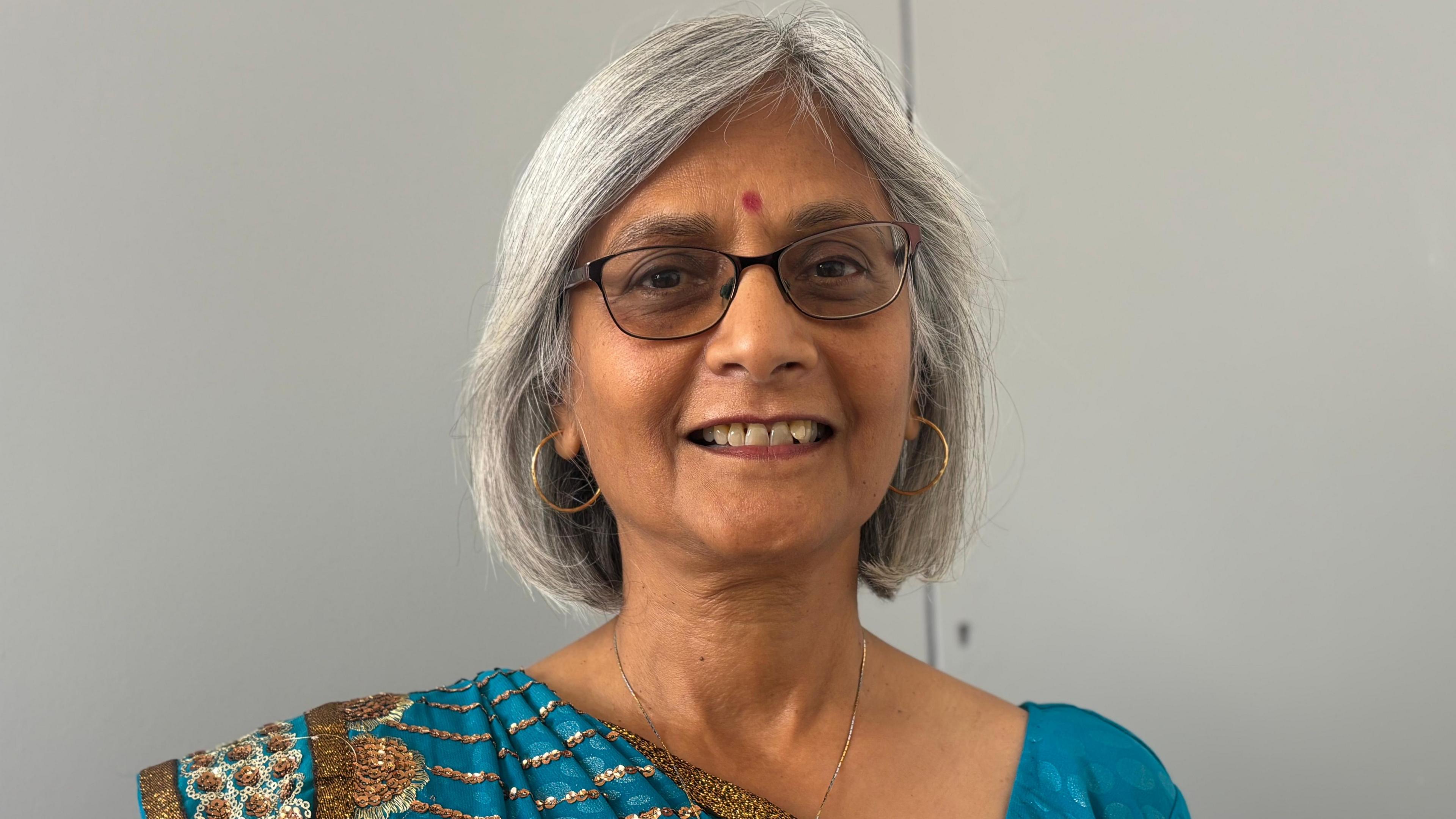 An Indian woman wearing a blue, white and brown saree. She is smiling at the camera and is wearing glasses and some earrings against a simple serene grey background. She has shoulder length long, dark hair with touches of grey. She has a red dot on her forehead, called "bindi," which is a traditional adornment in South Asian cultures, particularly Hinduism.