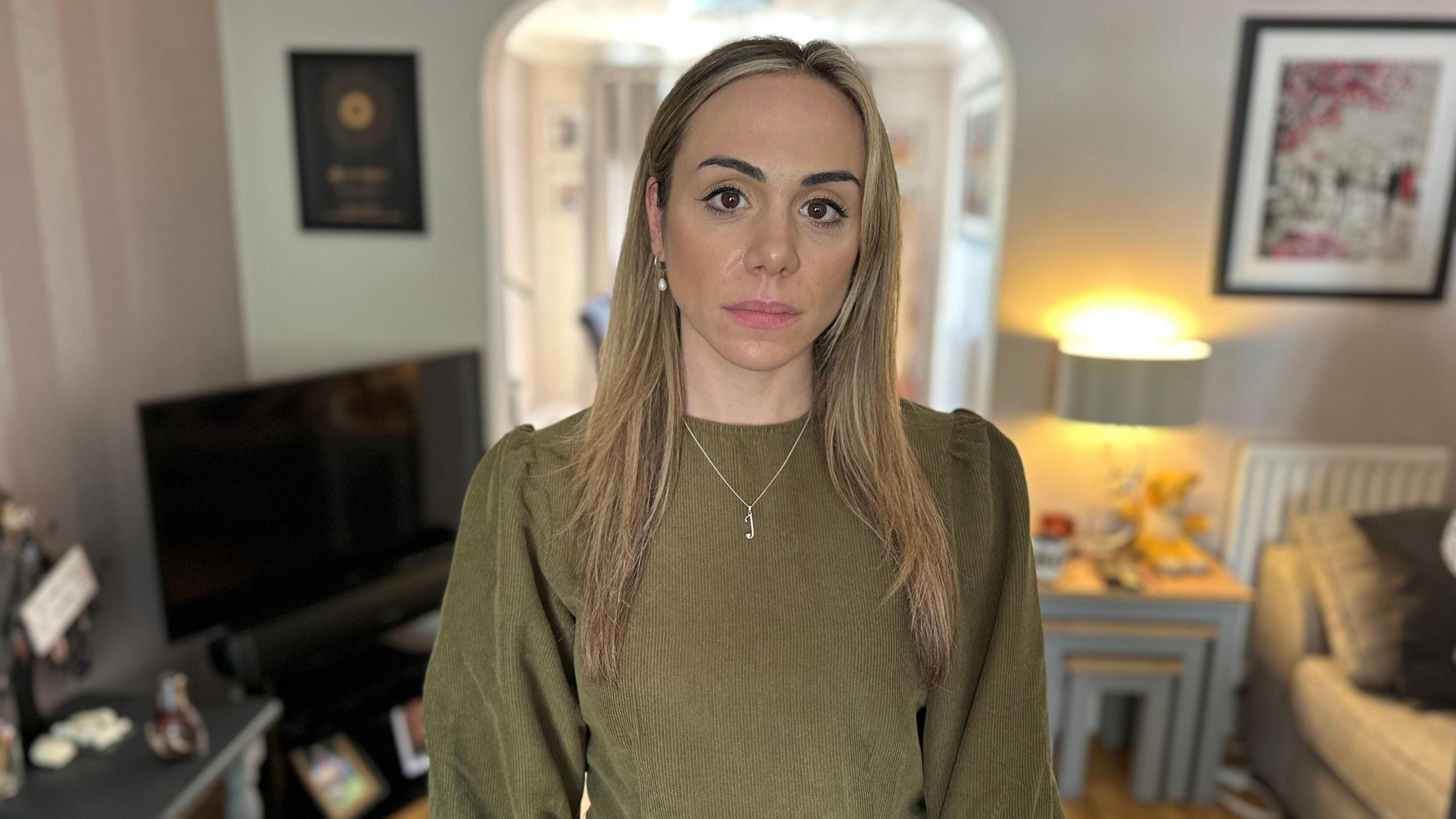 Mimi has long light brown hair and brown eyes. She is wearing a long-sleeced brown dress and is standing in her living room.