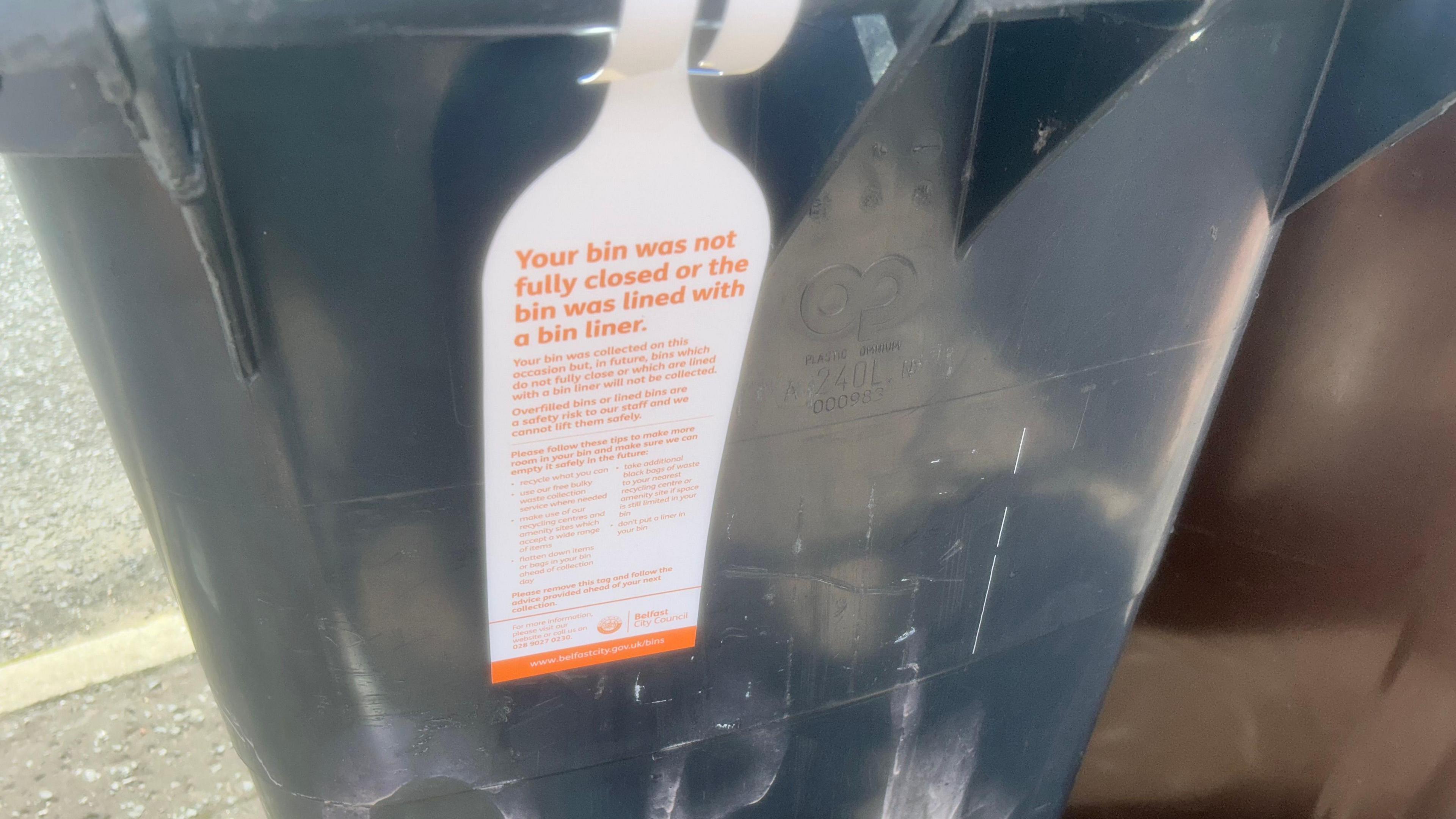 A white label, with orange writing hangs from a black bin. The text says 'Your bin was not fully closed or the bin was lined with a bin liner.' There is further, smaller writing on the label. There is also a Belfast city council logo at the bottom.  