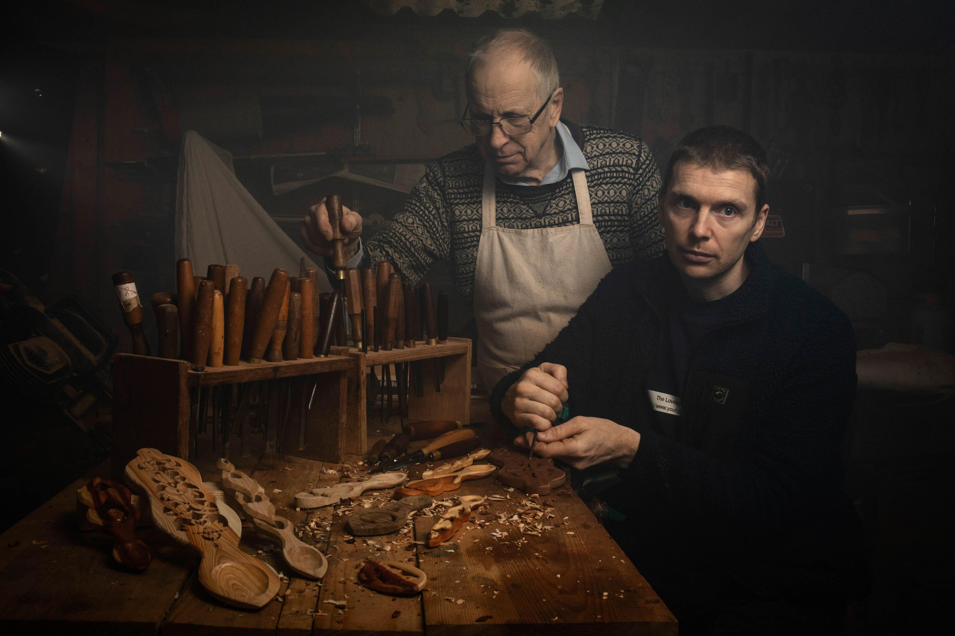 Two men at a carpentry table. One is sat down and is chipping something at the table. He looks at the camera with a vacant expression. He has short black hair and blue eyes. He wears a black cardigan. There is a man behind him with balding grey hair, he has a cream apron on with a green patterned jumper underneath and the collars of the blue shirt under that can be seen. There are various tools on the table and Welsh love spoons.