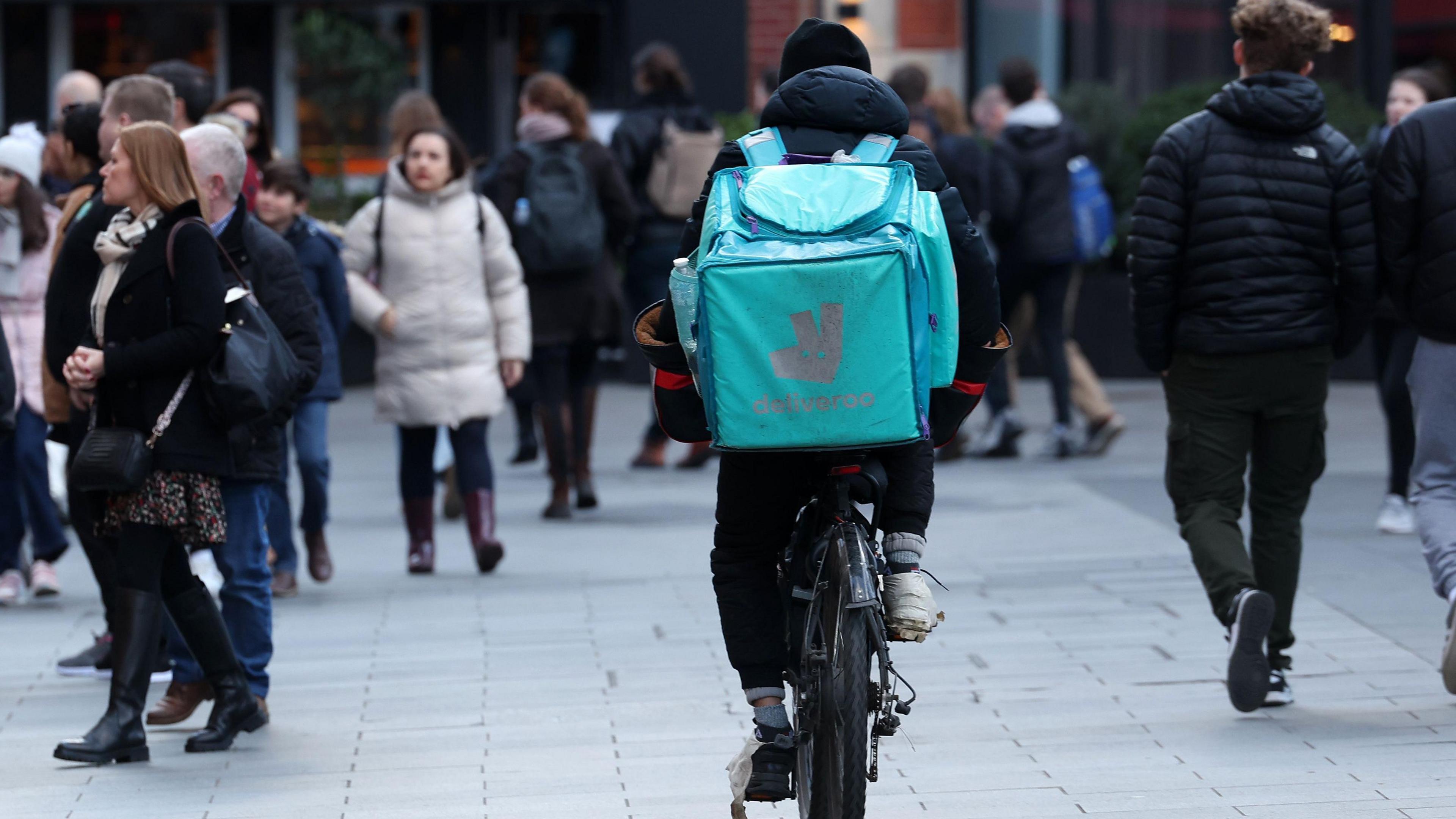 Deliveroo delivery driver rides a bicycle in London, Britain, 12 February 2024 - pictured from behind he is moving through and area crowded with pedestrians with his food delivery bag clearly visible on his back