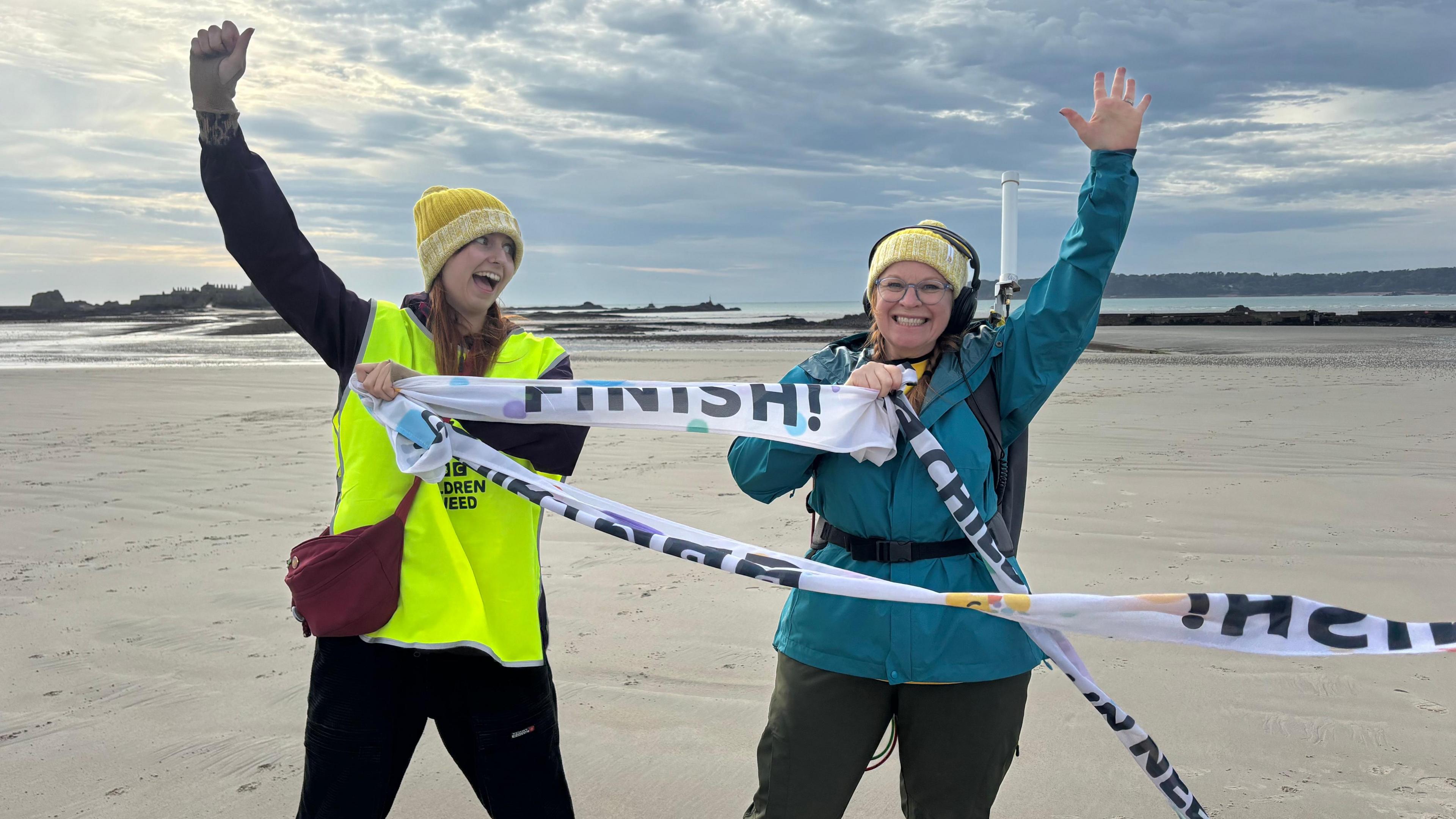 A picture of Alison Moss with Frankie Golding. Alison on the right, is wearing a blue coat and holding the finish tape. Frankie on the left, is wearing a green and hat.