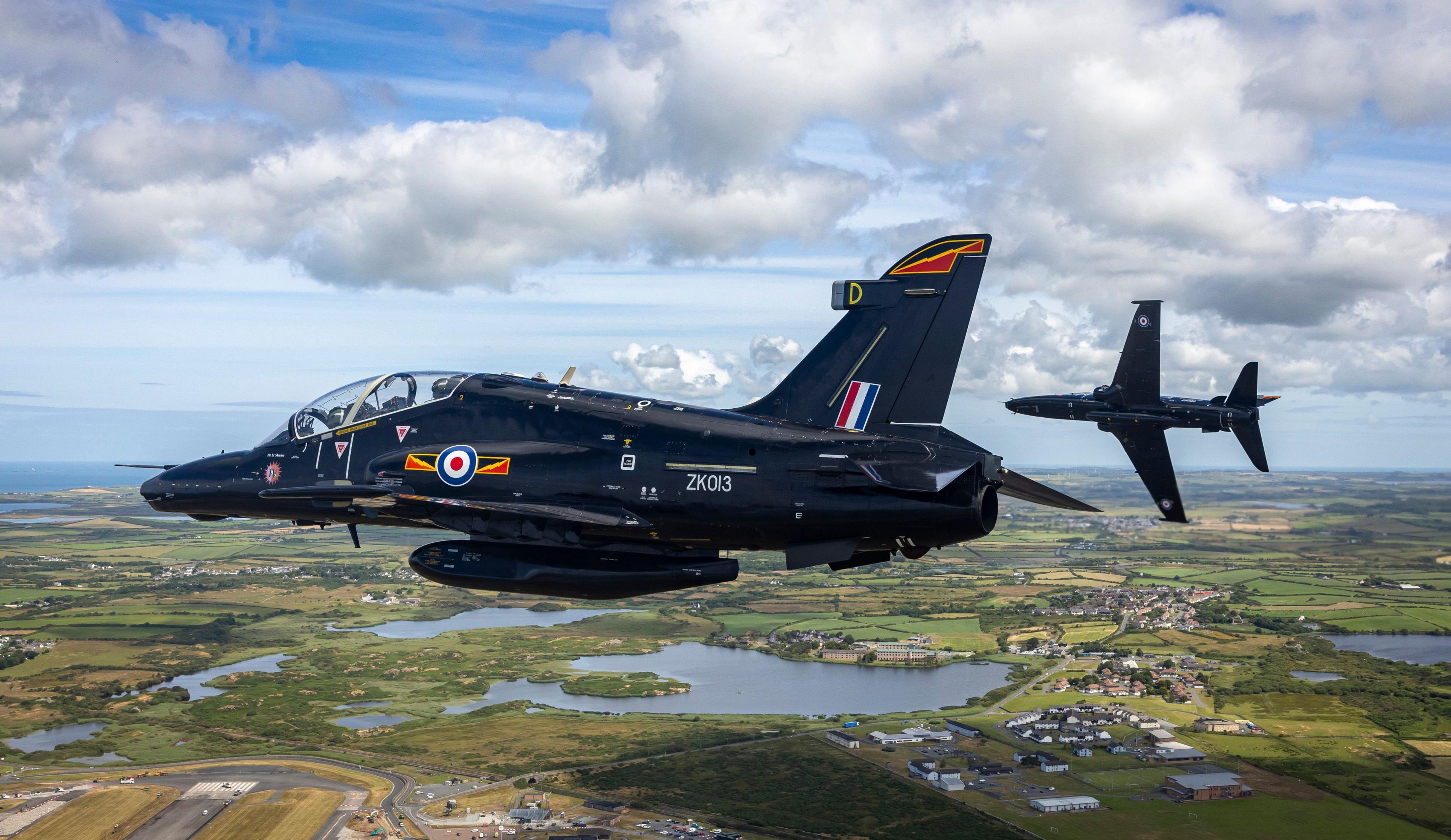 Two navy jet planes seen in the air with fields and lakes below in blue skies with clouds