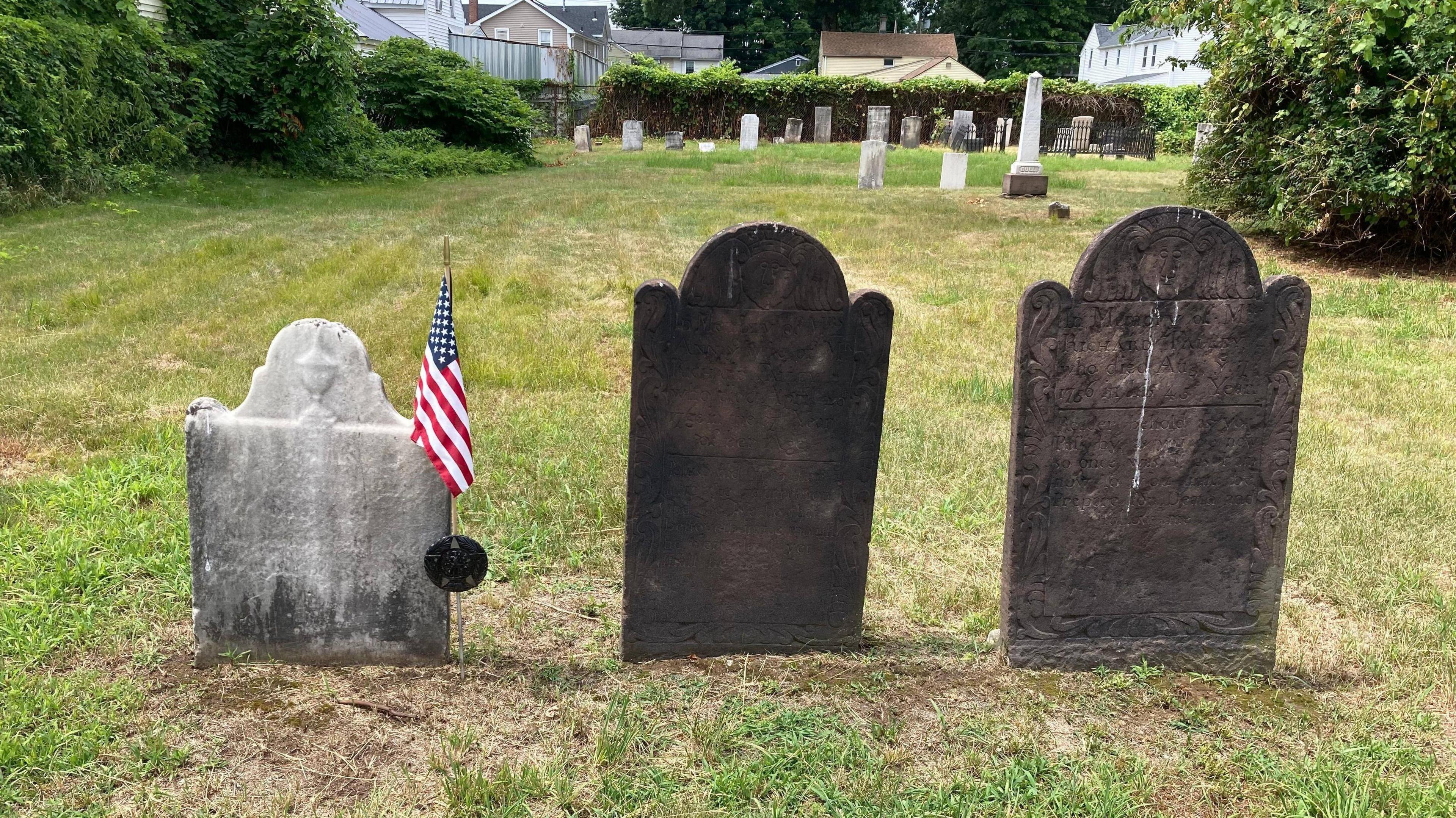 Three gravestones in a grassy cemetery. One is made of white stone and has a US flag next to it, while the others are made of darker, almost black stone.