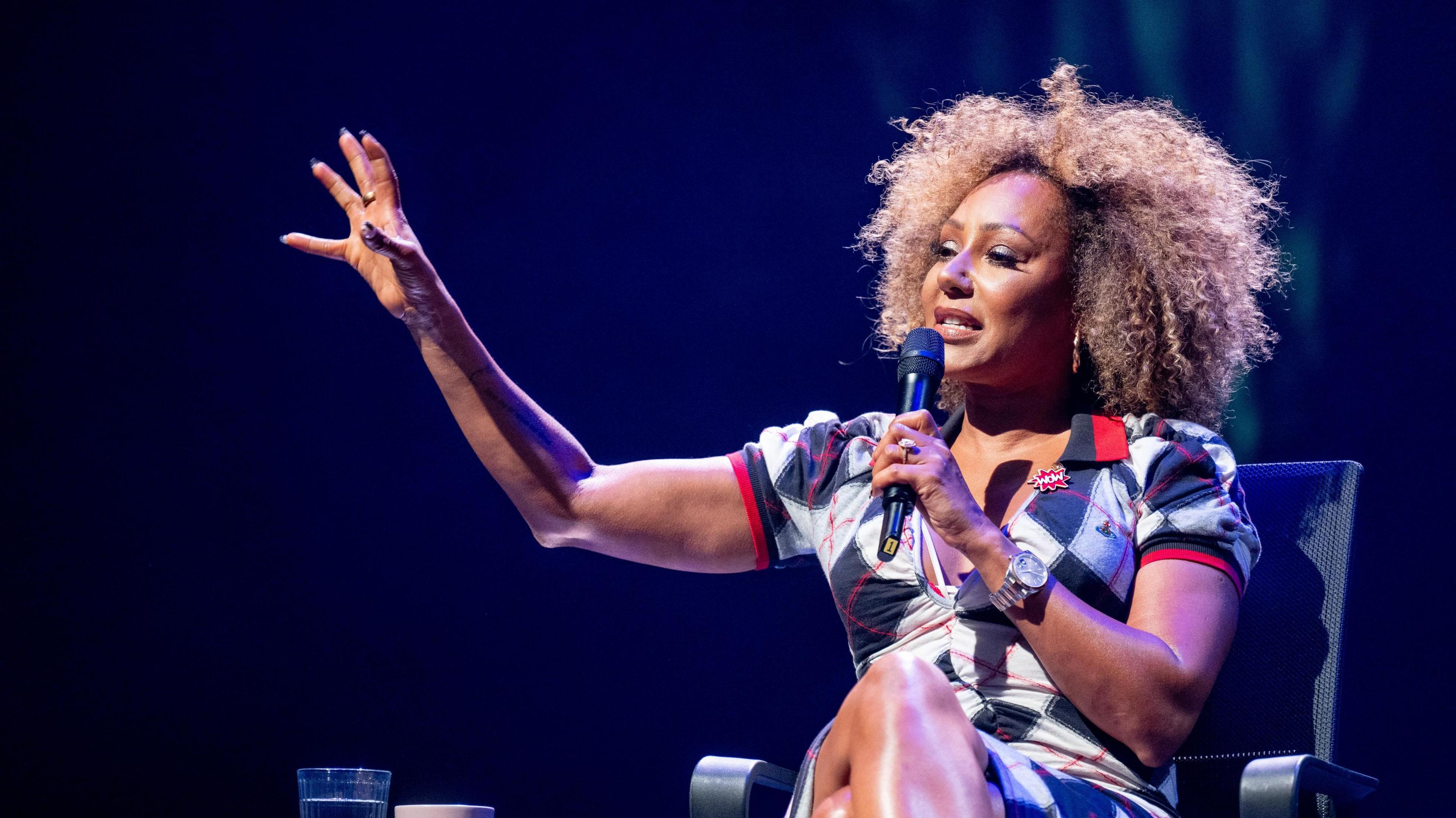 Mel B sat on a black chair with a microphone to her mouth, in what appears to be a theatre. 