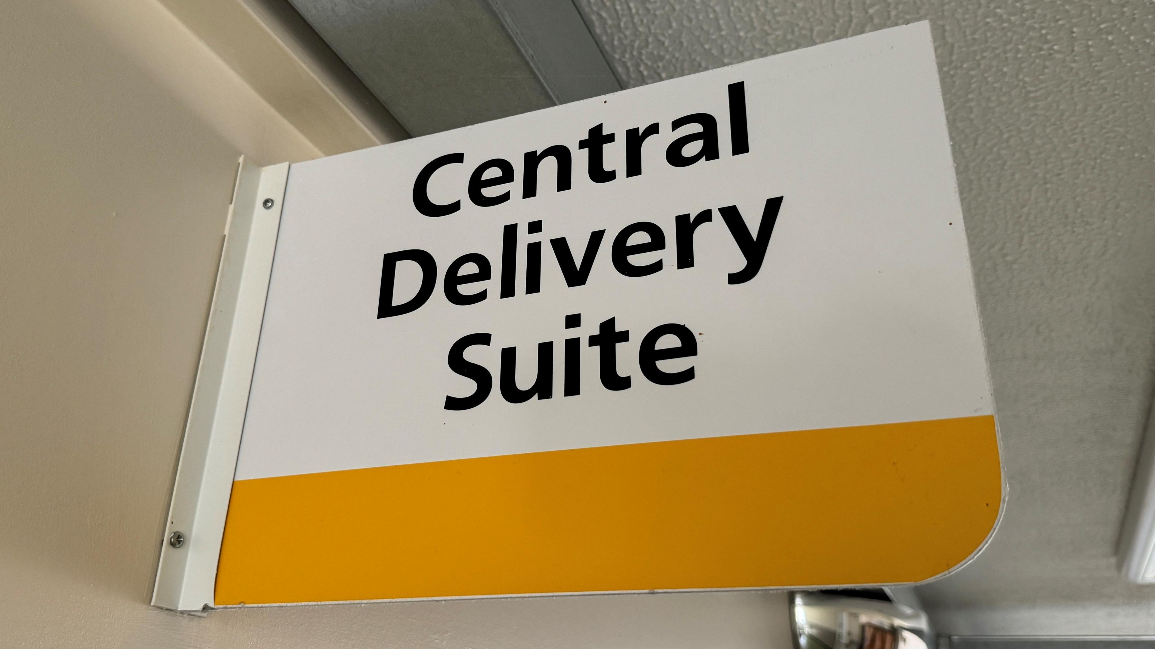 A white sign with black writing saying Central Delivery Suite. There is a blank yellow strip underneath the writing for design purposes.