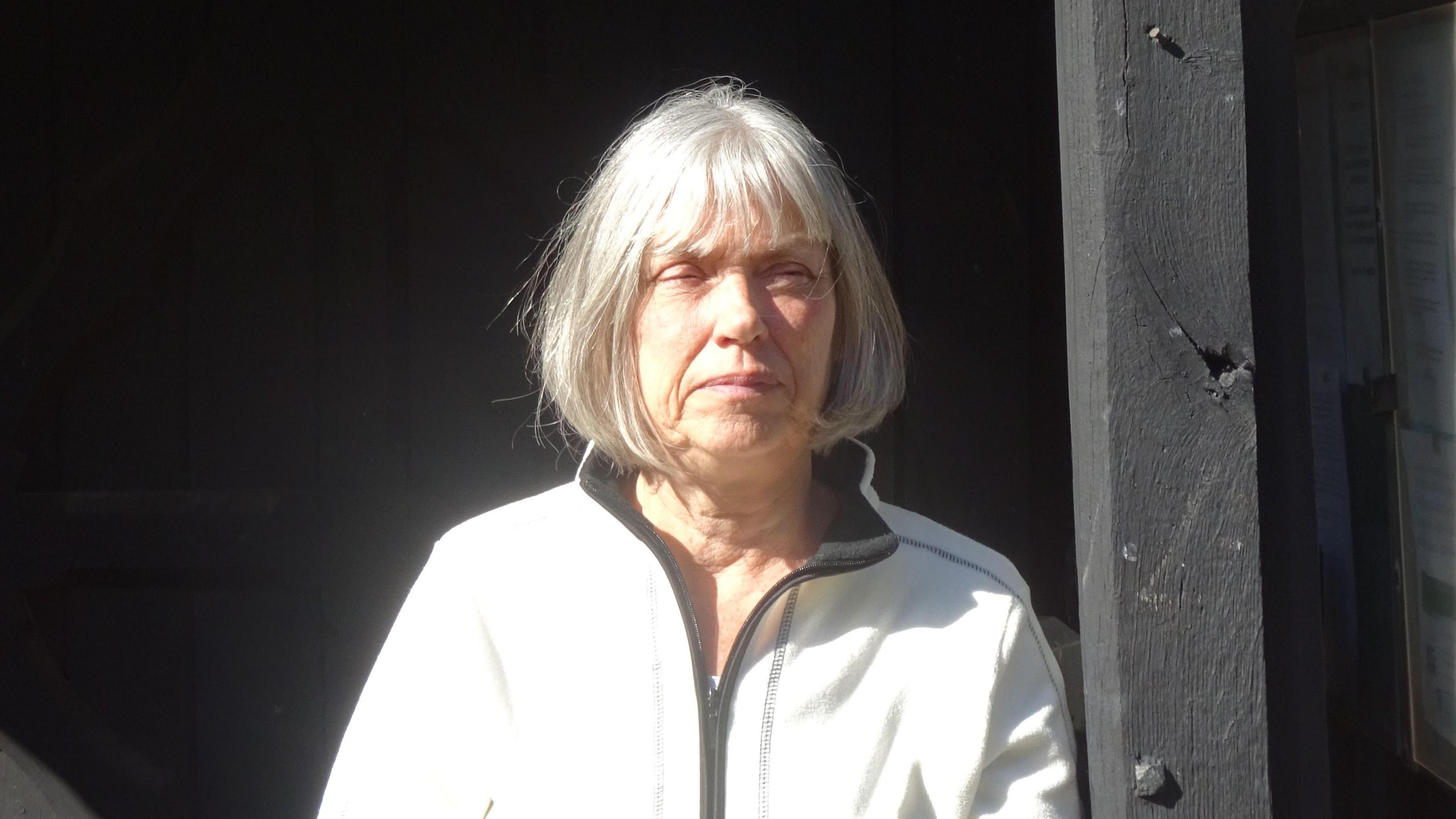 Jacquie Eykelbosch, leaning against a black timber upright. She has bobbed grey hair and is wearing a white top with a black zip. She is squinting slightly in the sun.