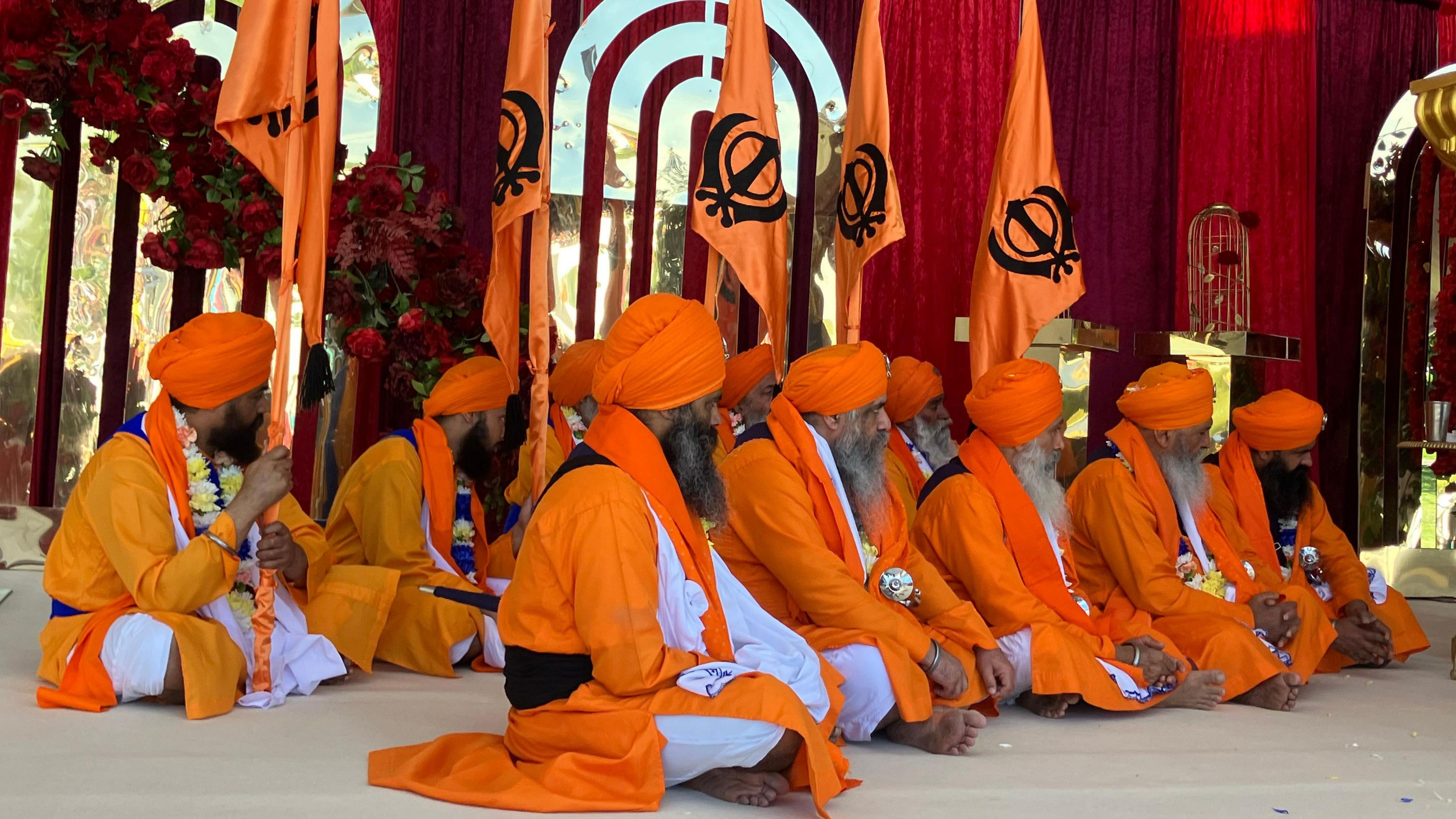 A group of men sitting cross-legged on the floor. They are wearing orange turbans and orange and white robes.