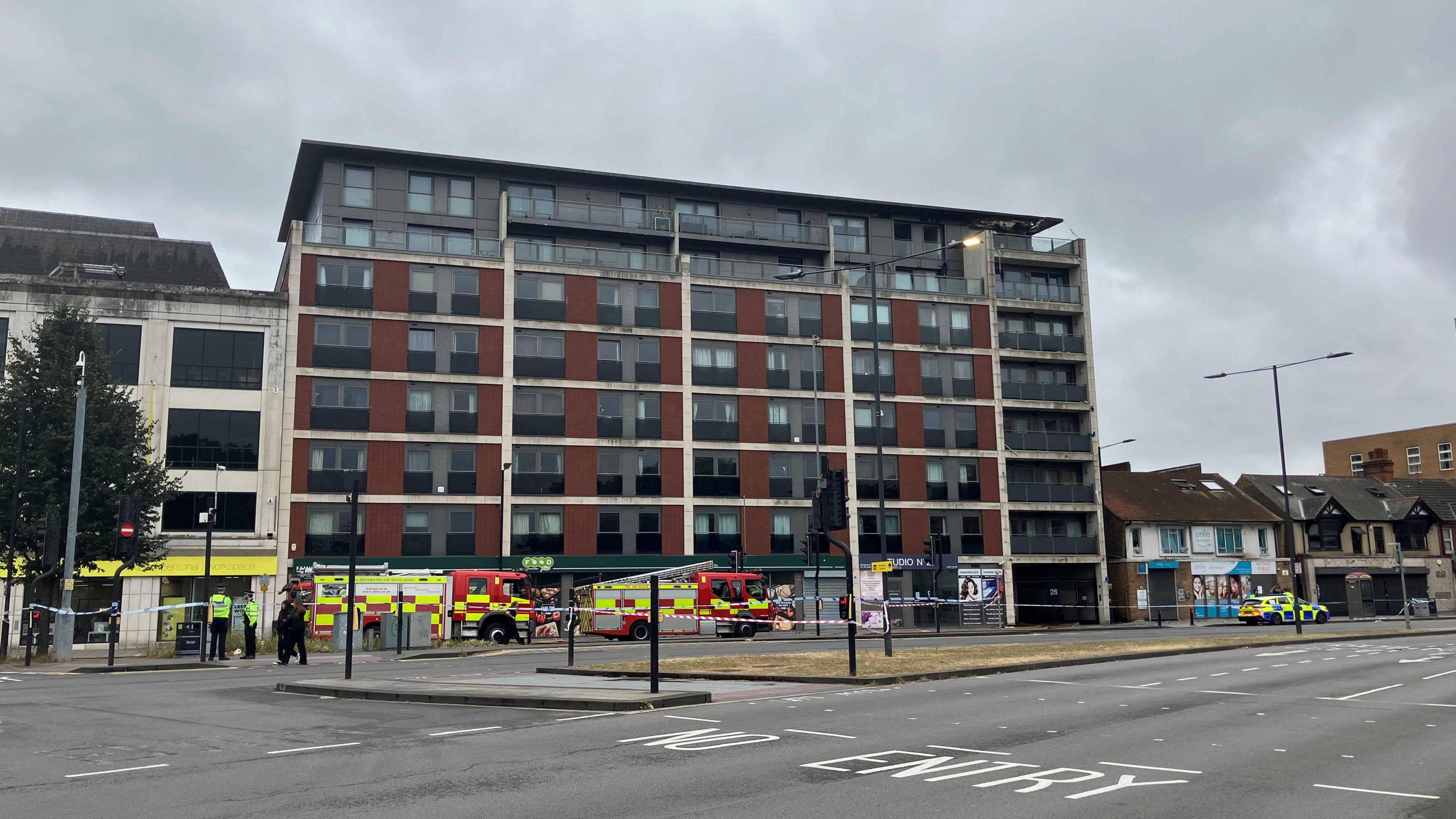 The red and grey apartment block has seven floors, with two fire trucks outside and a police car. The seventh and sixth floor been burnt and whole building evacuated. 