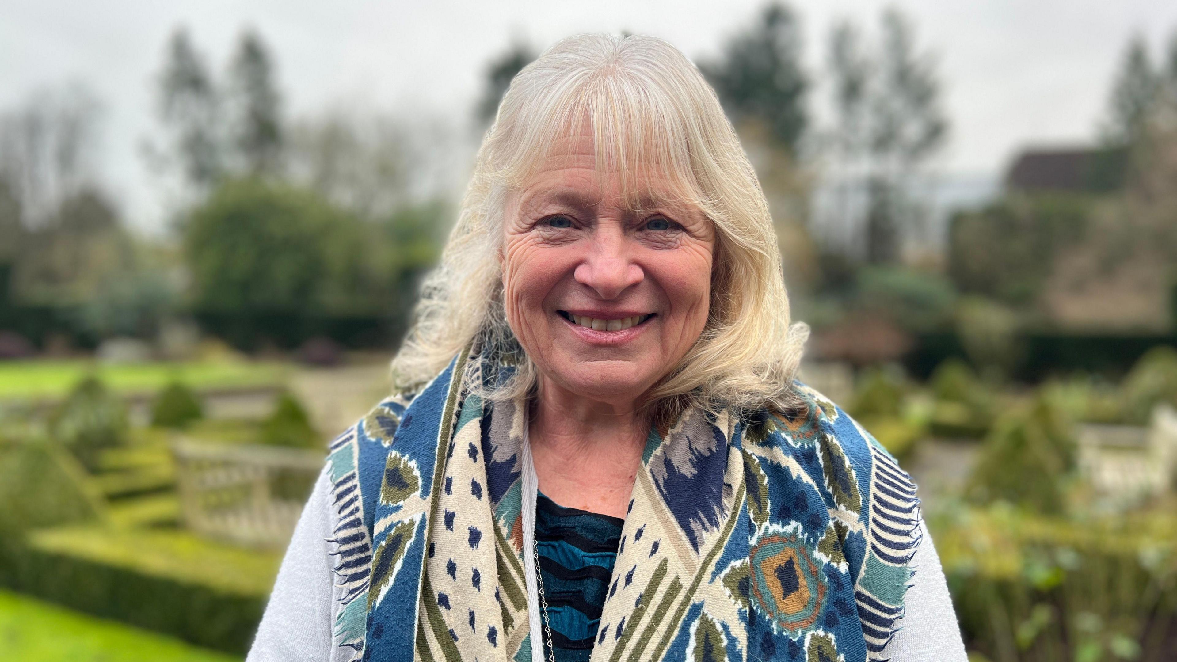 Anne Swithinbank is seen from her shoulders up. She is wearing a blue, patterned scarf and has light, shoulder-length hair. She is smiling. In the background is a pristine garden at a country house.