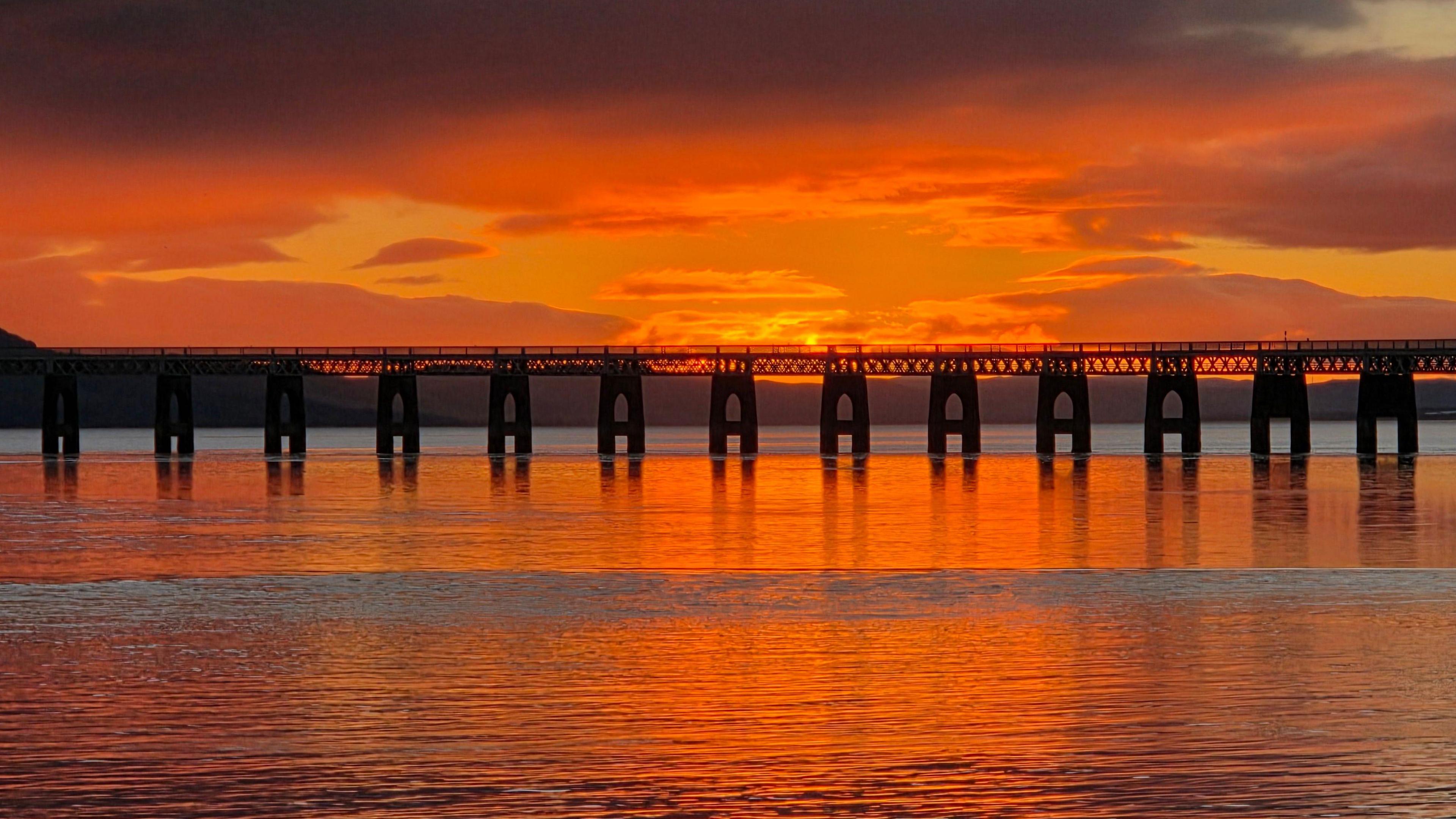 A long bridge stretches across calm water under a fiery orange sunset sky.