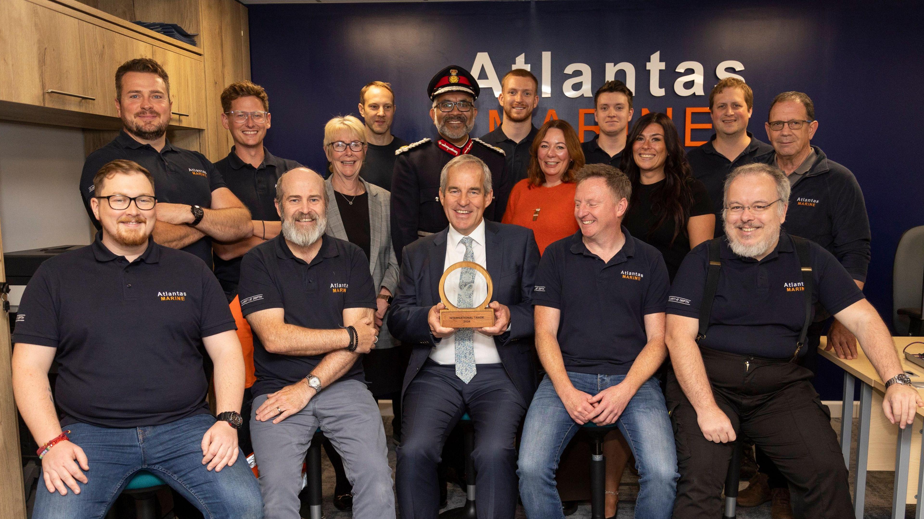 Charlie Foll from Atlantas Marine sits alongside team members whilst holding King's Award presented to him by Lord-Lieutenant of Somerset who is stood behind him.