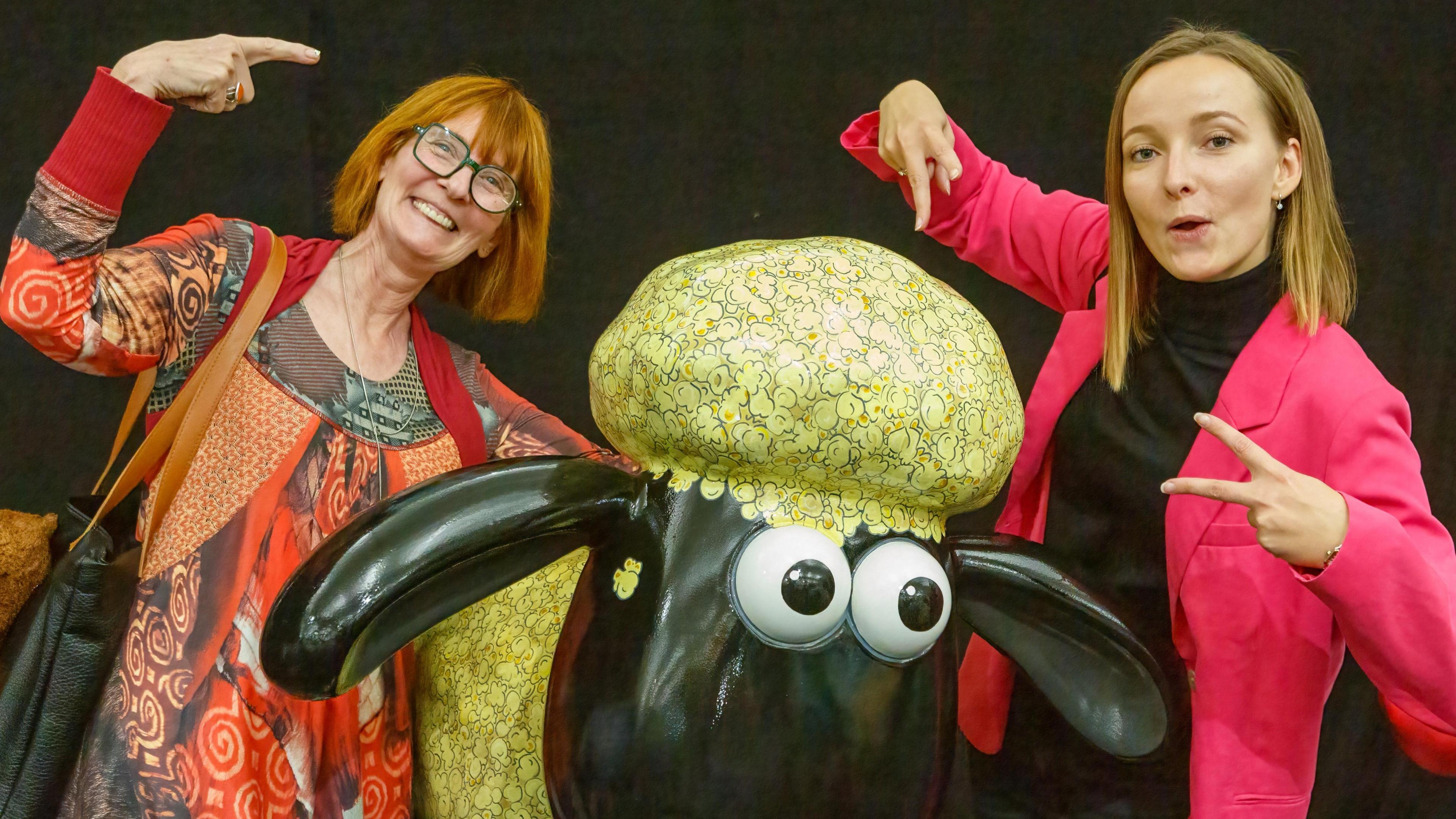 Two women stand next to a Shaun the Sheep statue. The lady on the left has a red bob and is wearing a red and orange patterned dress. She is doing an Ali G style hand gesture. The lady on the right has blonde hair and is wearing a pink blazer with a black top. She is doing peace signs. 