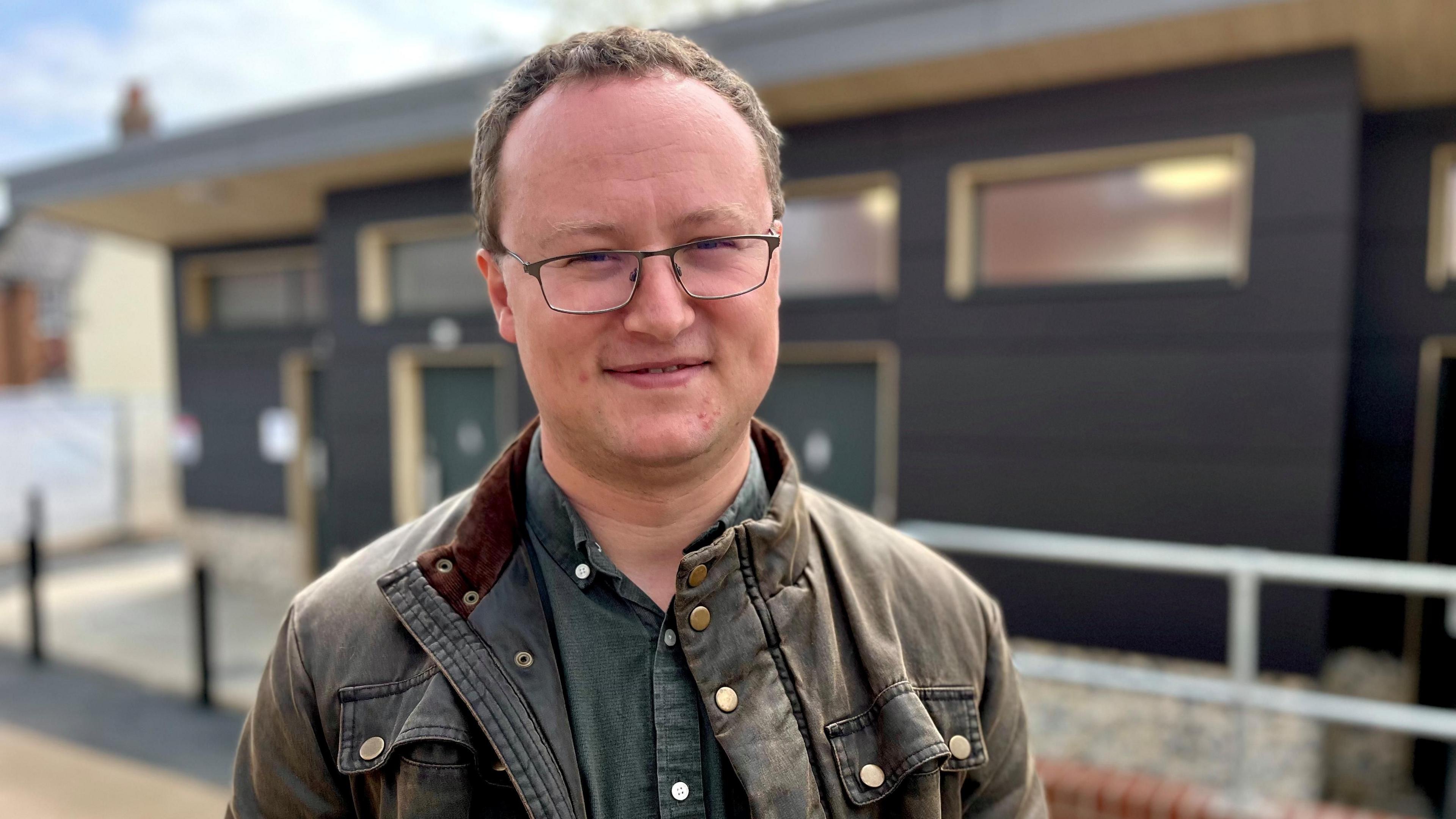 Tim Adams has short brown hair and is wearing glasses. He is wearing a brown jacket with a green shirt underneath.
He is smiling at the camera. A toilet block is behind him.