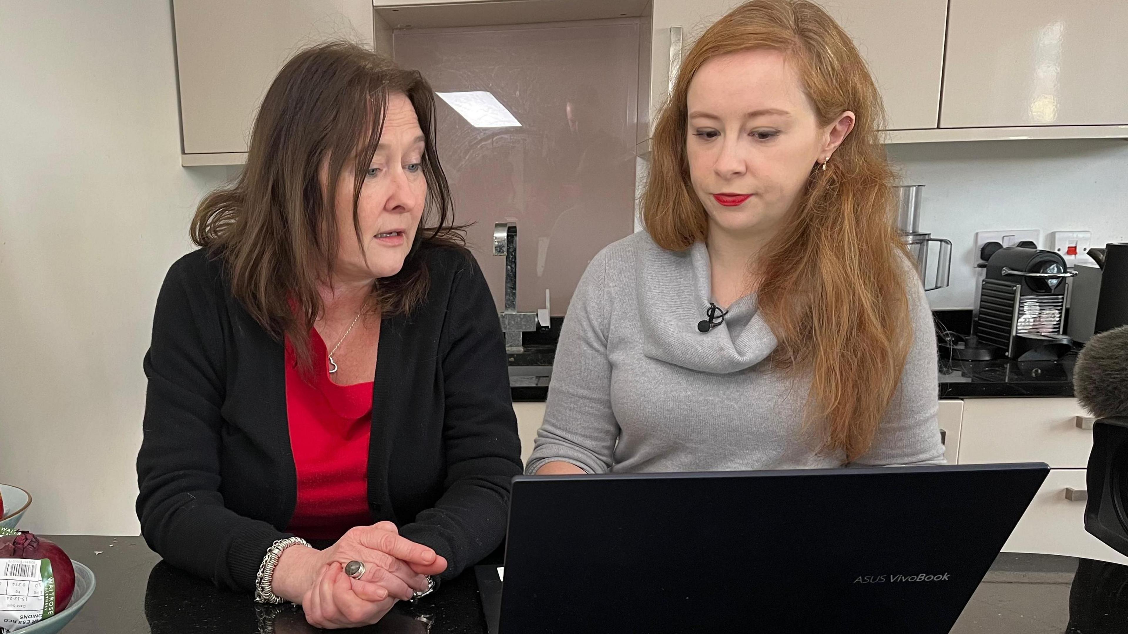 Two women are seated at a black countertop in a kitchen. One woman with dark hair is wearing a red top and black cardigan, and the other, with long red hair, is wearing a light grey top and using a laptop.