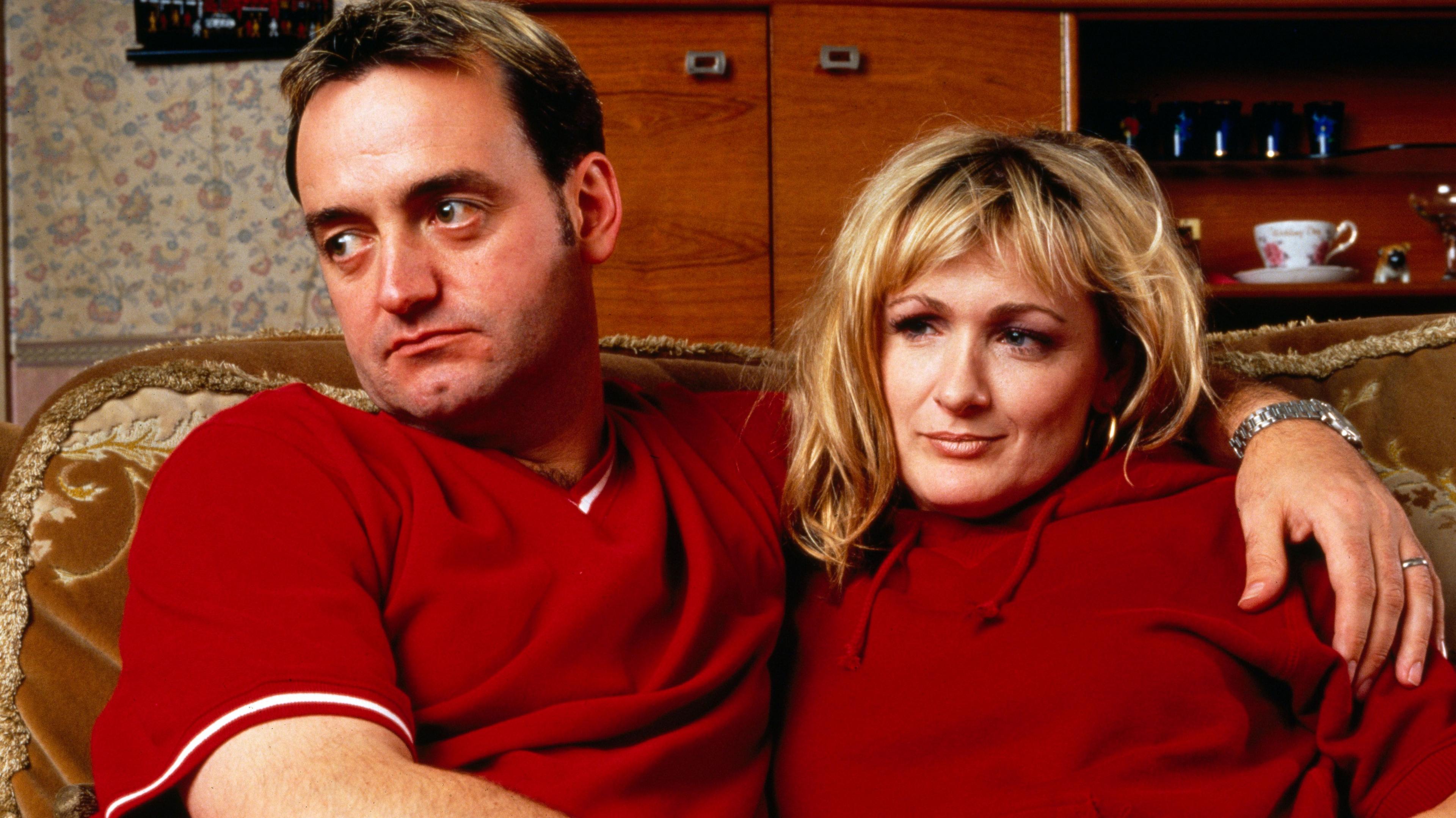 Craig Cash and Caroline Aherne, as Dave and Denise from The Royle Family, both dressed in red and sitting on the sofa from the Royle living room.