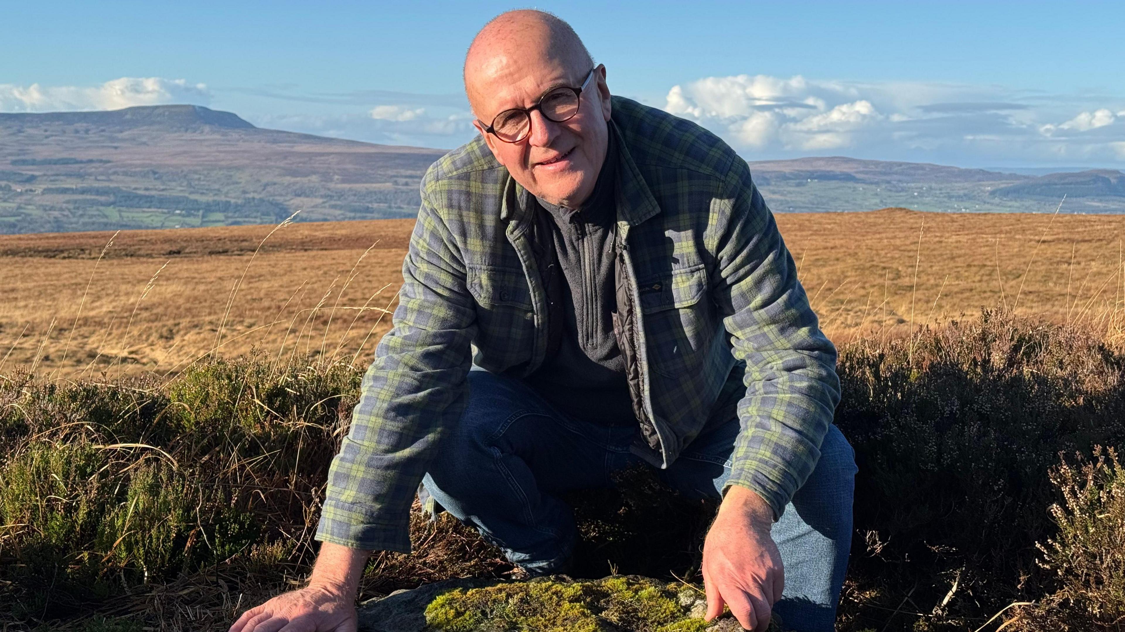 Barney Devine crouches over a stone in the middle of a grass plane with some hills in the background. He is bald, wears glasses and a green checked shirt and light blue jeans and he is looking at the camera.