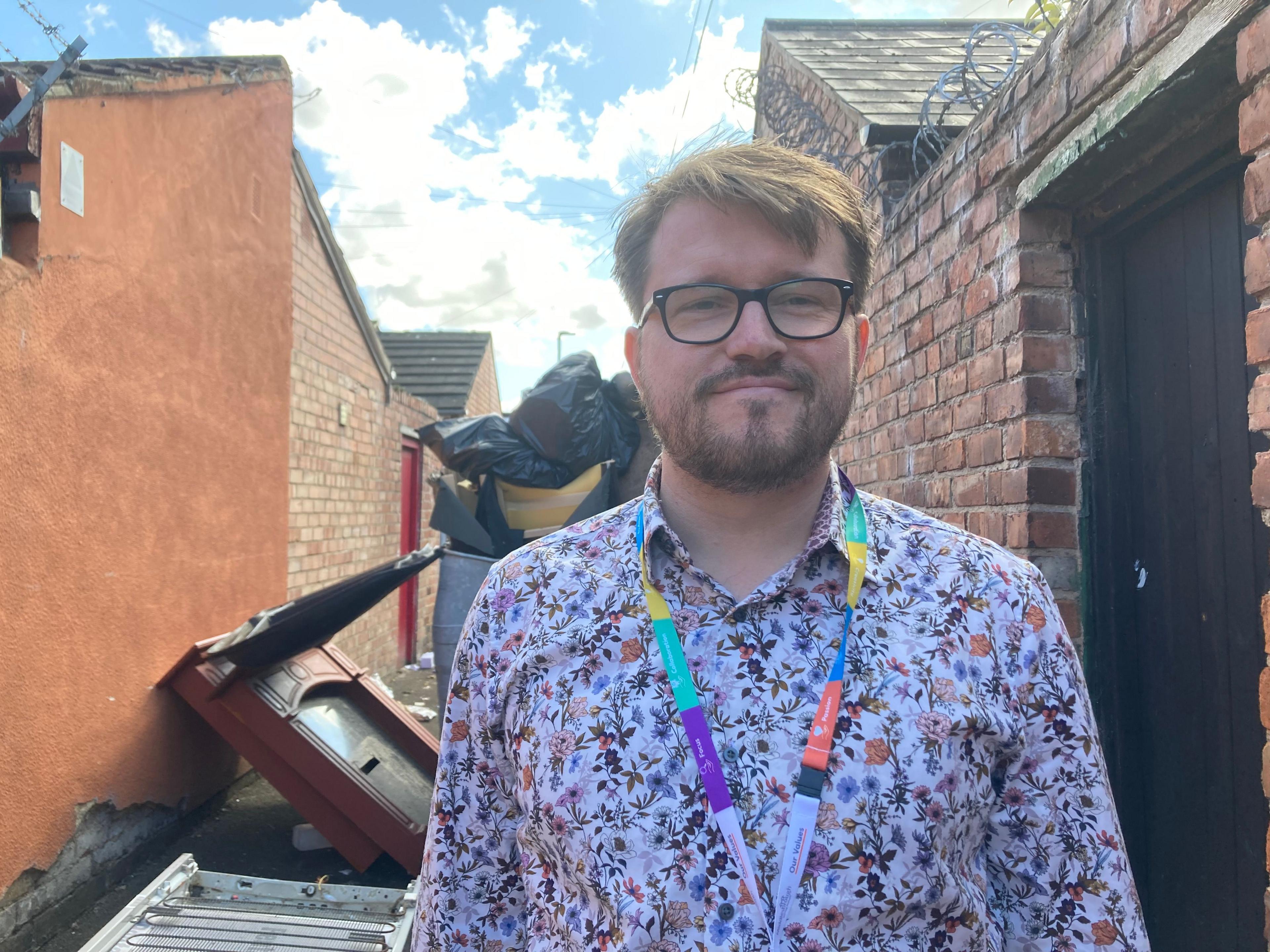  Lewis Young stands in front of the rubbish bags dumped in an alley. He has short brown hair and wears a flowery-patterned shirt with a multi-coloured lanyard. An old fireplace is behind him.