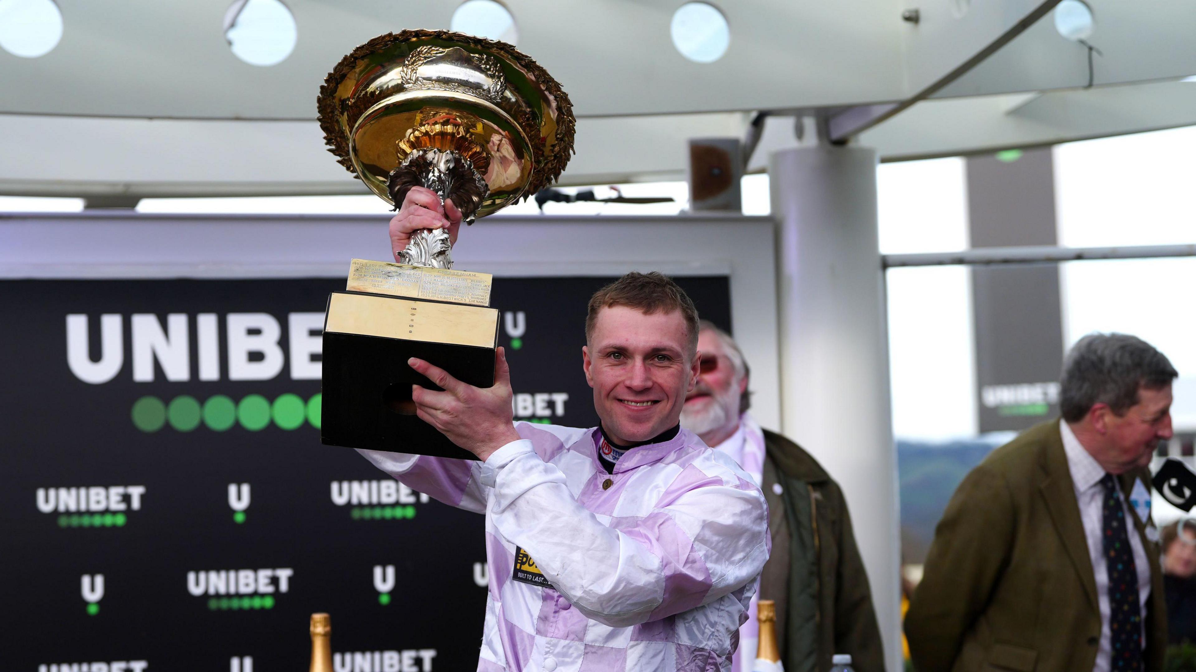 Lorcan Williams smiles as he holds up a large trophy with two arms. He wears a lilac and white checked racing shirt.